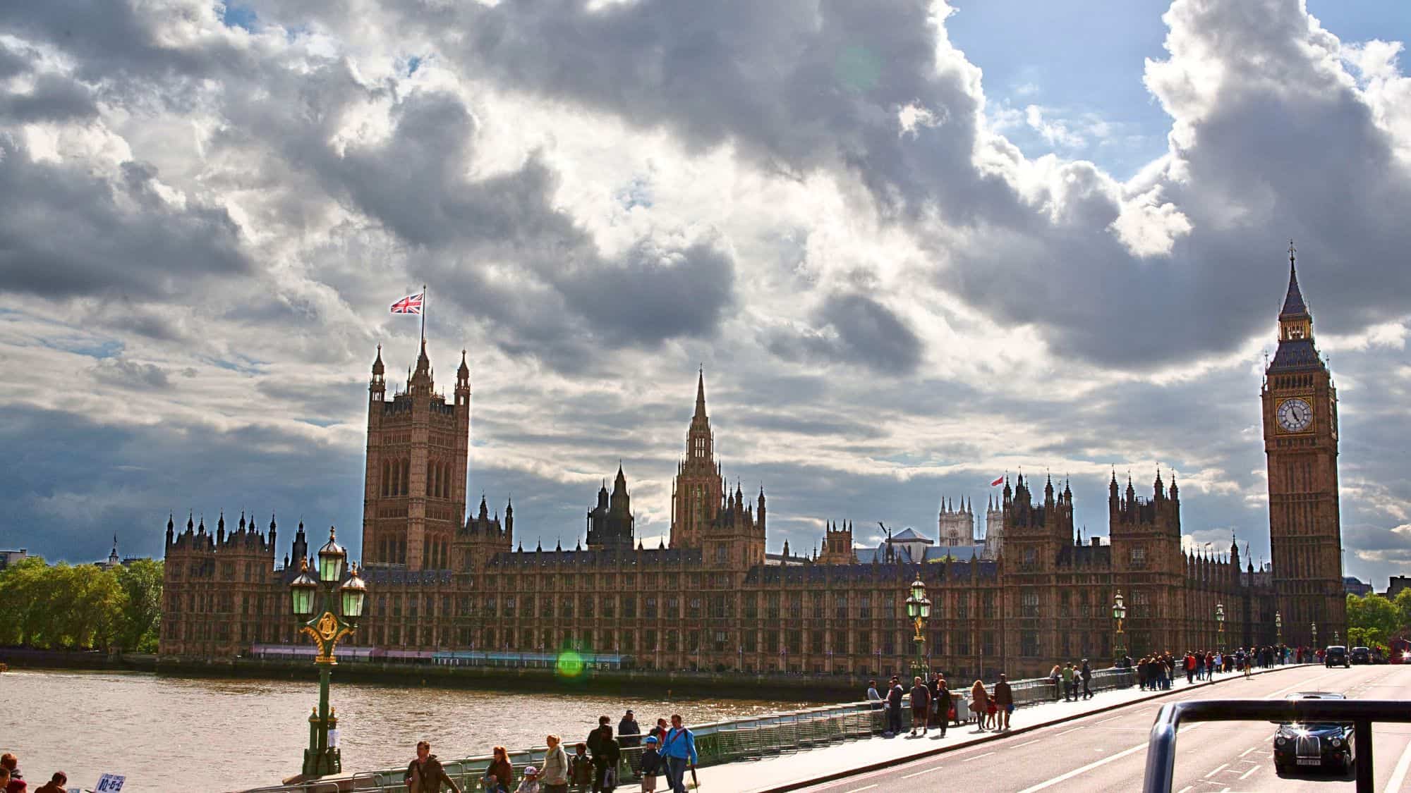 The iconic Palace of Westminster, home to the UK Parliament, sits along the River Thames. Big Ben towers to the right, while the Union Jack flies under a partly cloudy sky.