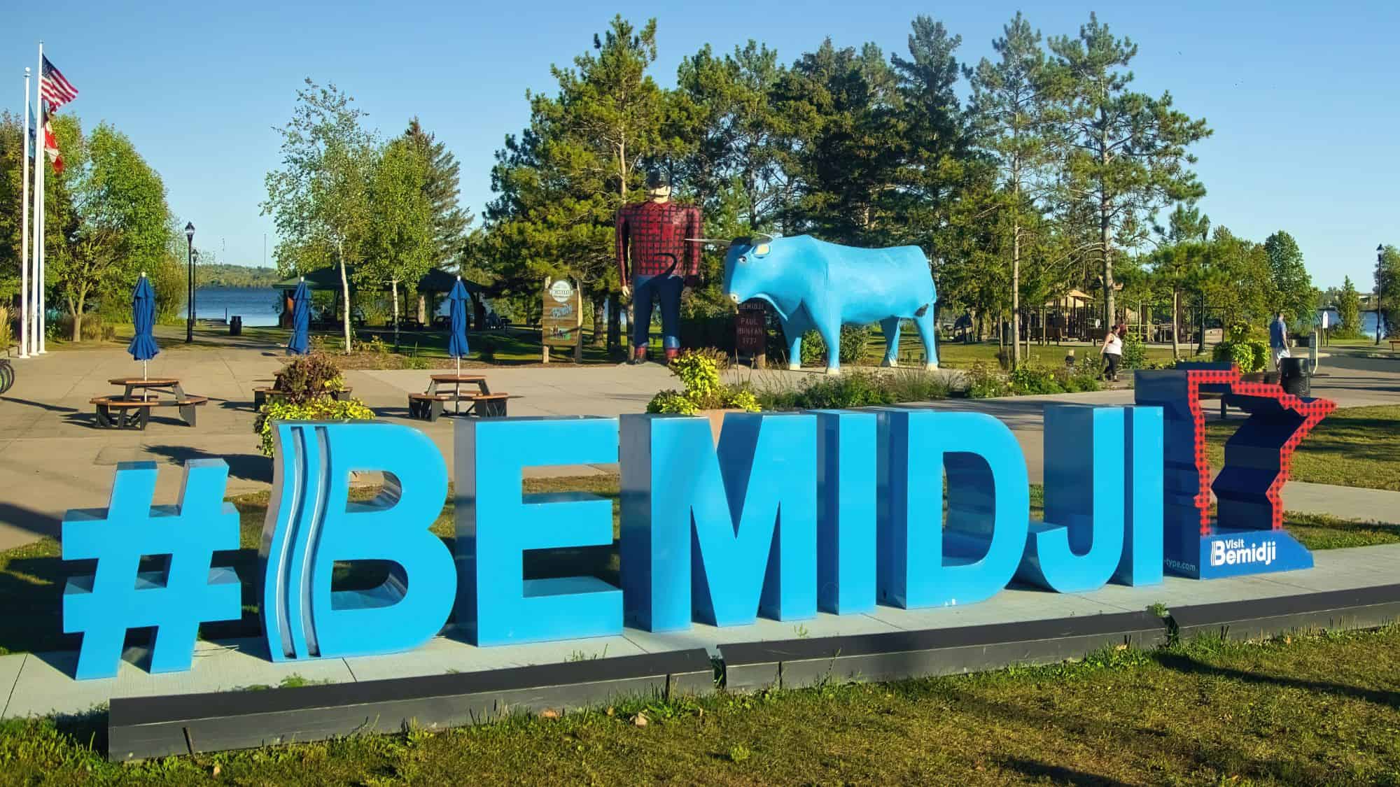 A vibrant blue sign reading "#BEMIDJI" stands in front of the iconic Paul Bunyan and Babe the Blue Ox statues in Bemidji, Minnesota. The oversized lumberjack and his blue ox are set against a scenic lakeside park with picnic tables and American and Canadian flags.