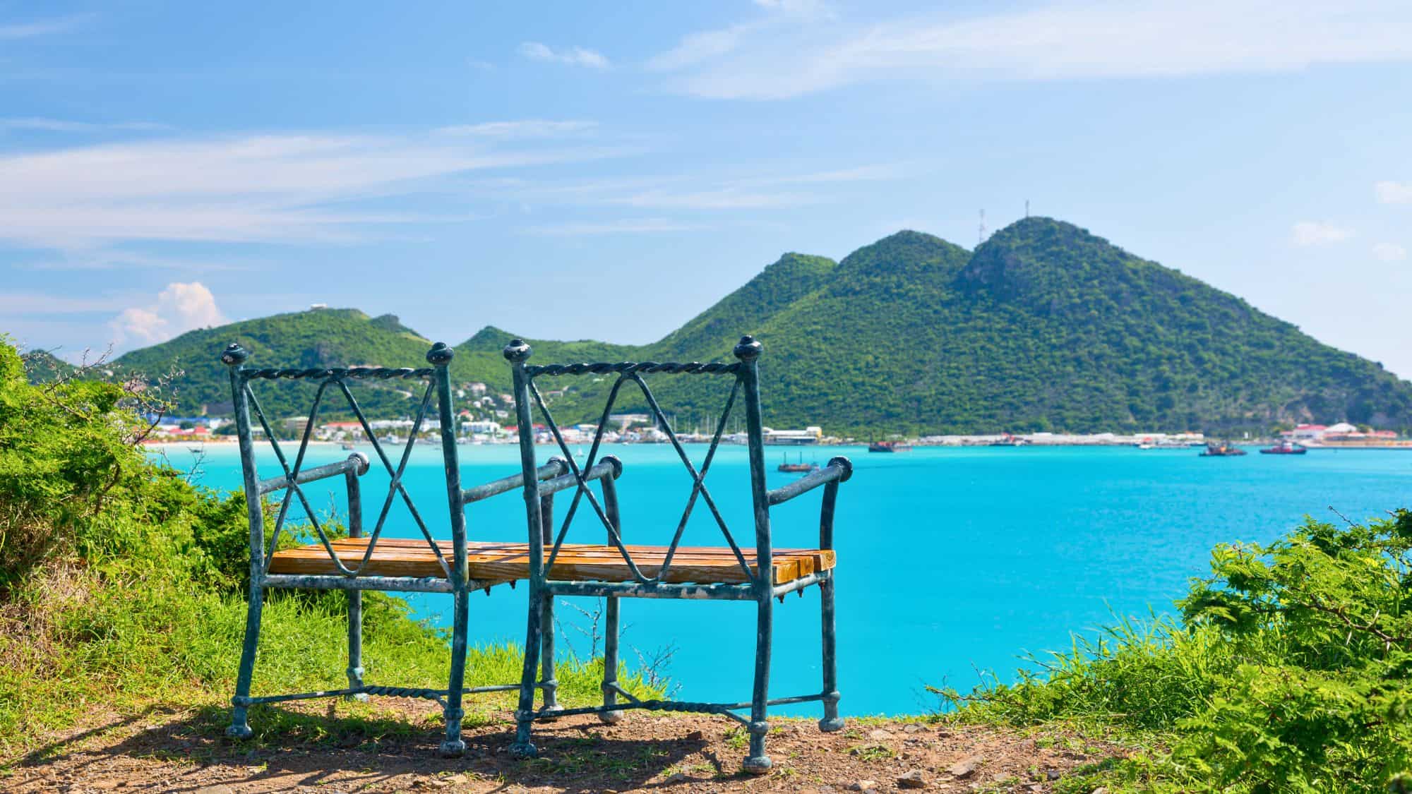 Two iron benches overlook a vibrant blue bay with lush green mountains in the background. The picturesque scene showcases boats in the harbor and colorful buildings along the shoreline.