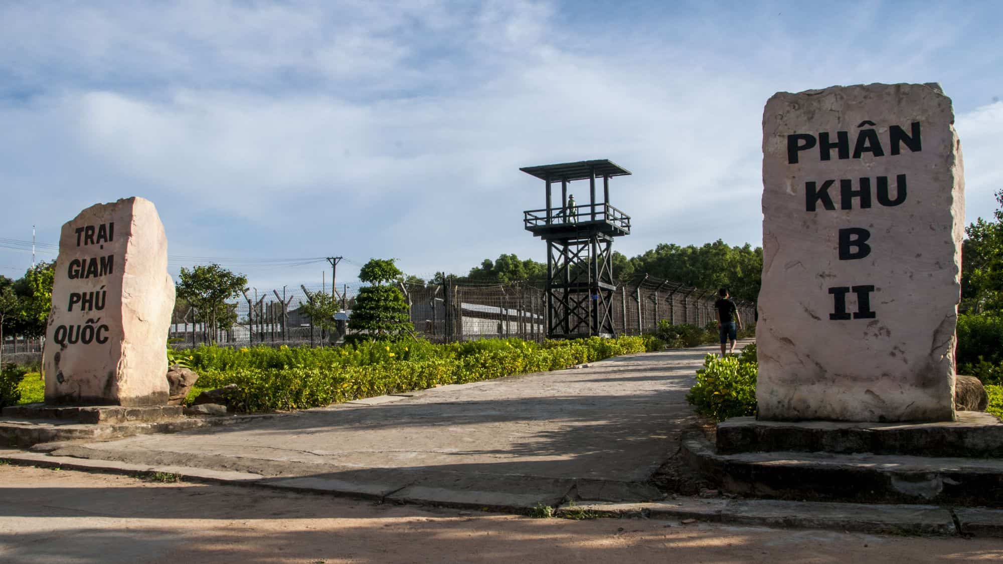 A historical site in Phu Quoc, Vietnam, showing large stone markers with Vietnamese inscriptions at the entrance, a watchtower in the background, and barbed wire fences surrounding the area.
