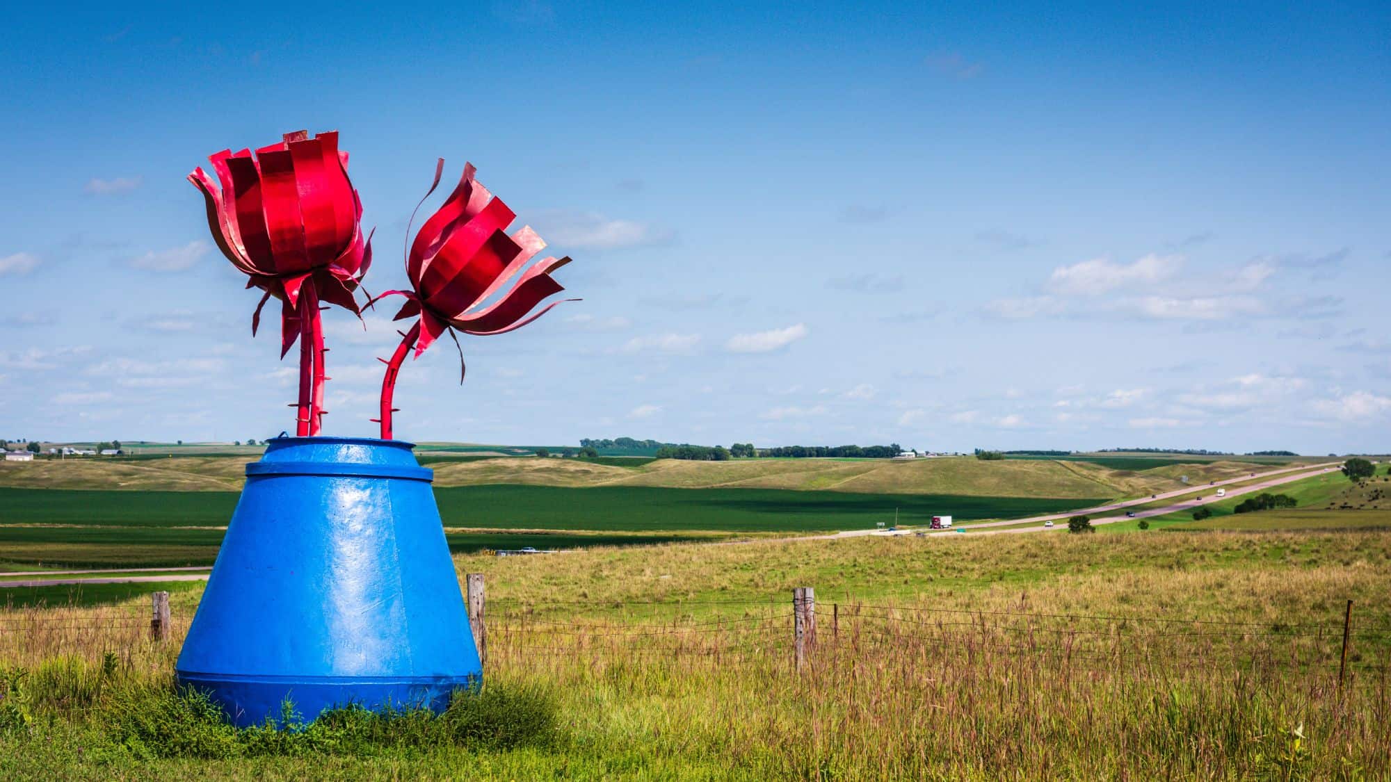 A roadside metal sculpture of two giant red tulips in a bright blue base stands against rolling green hills and a clear blue sky. The artwork is part of the Porter Sculpture Park, known for its whimsical, oversized sculptures.