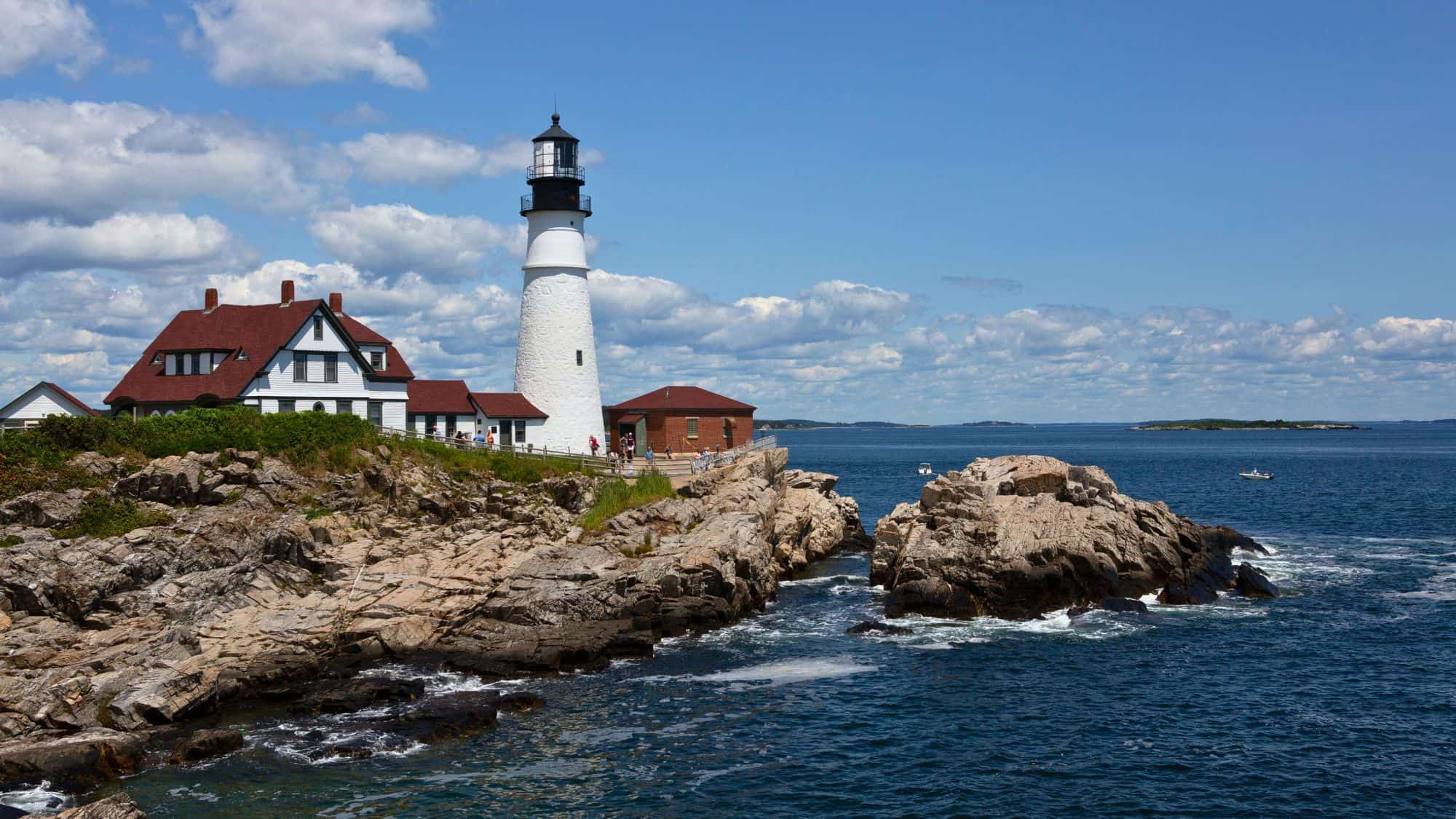 A classic New England lighthouse perched on rocky cliffs, overlooking the deep blue ocean. The white lighthouse with its red-roofed keeper’s house is surrounded by visitors taking in the coastal views.