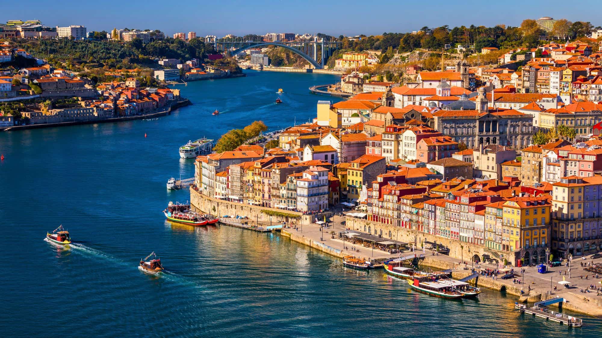 A panoramic view of Porto’s colorful buildings lining the Douro River, with boats cruising along the water. The city’s iconic red-tiled rooftops and historic architecture create a picturesque European charm.