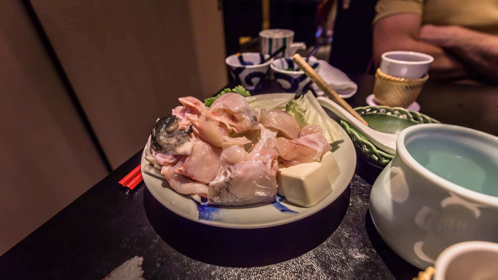 A plate of raw pufferfish sashimi with tofu and vegetables, ready for cooking. The setting includes traditional Japanese dishware and chopsticks, suggesting a fine dining experience.