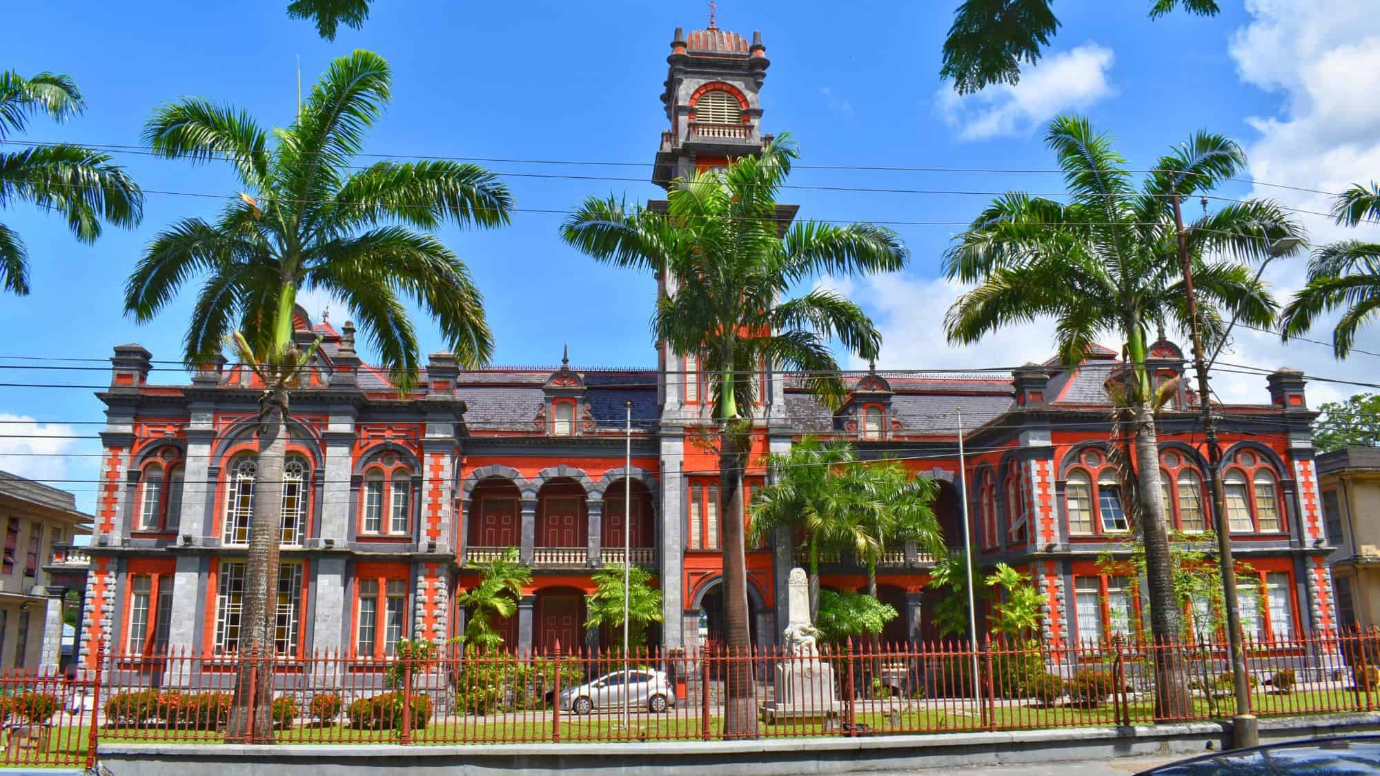 A grand colonial-style red and gray building with arched windows and a central clock tower, framed by tall palm trees, stands in Queen’s Park Savannah in Port of Spain, Trinidad and Tobago.