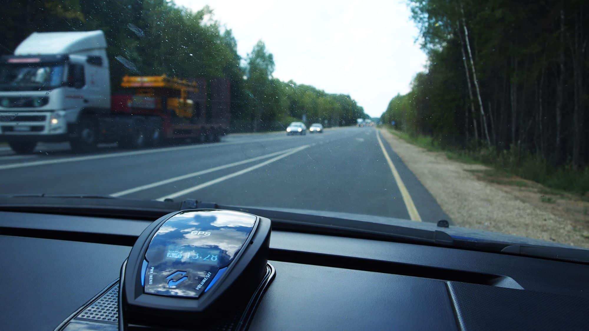 A dashboard view of a car with a radar detector mounted near the windshield. The road ahead is visible with trucks and cars in the distance.