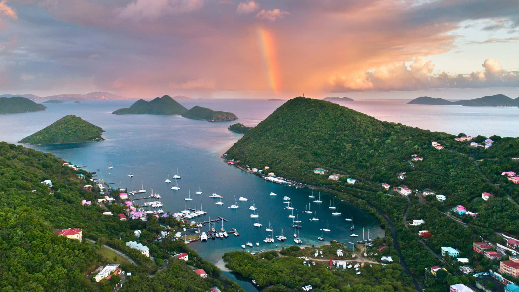 A serene harbor dotted with sailboats, surrounded by lush green hills and quaint houses. A rainbow arches over the distant islands under a dramatic, colorful sunset sky.