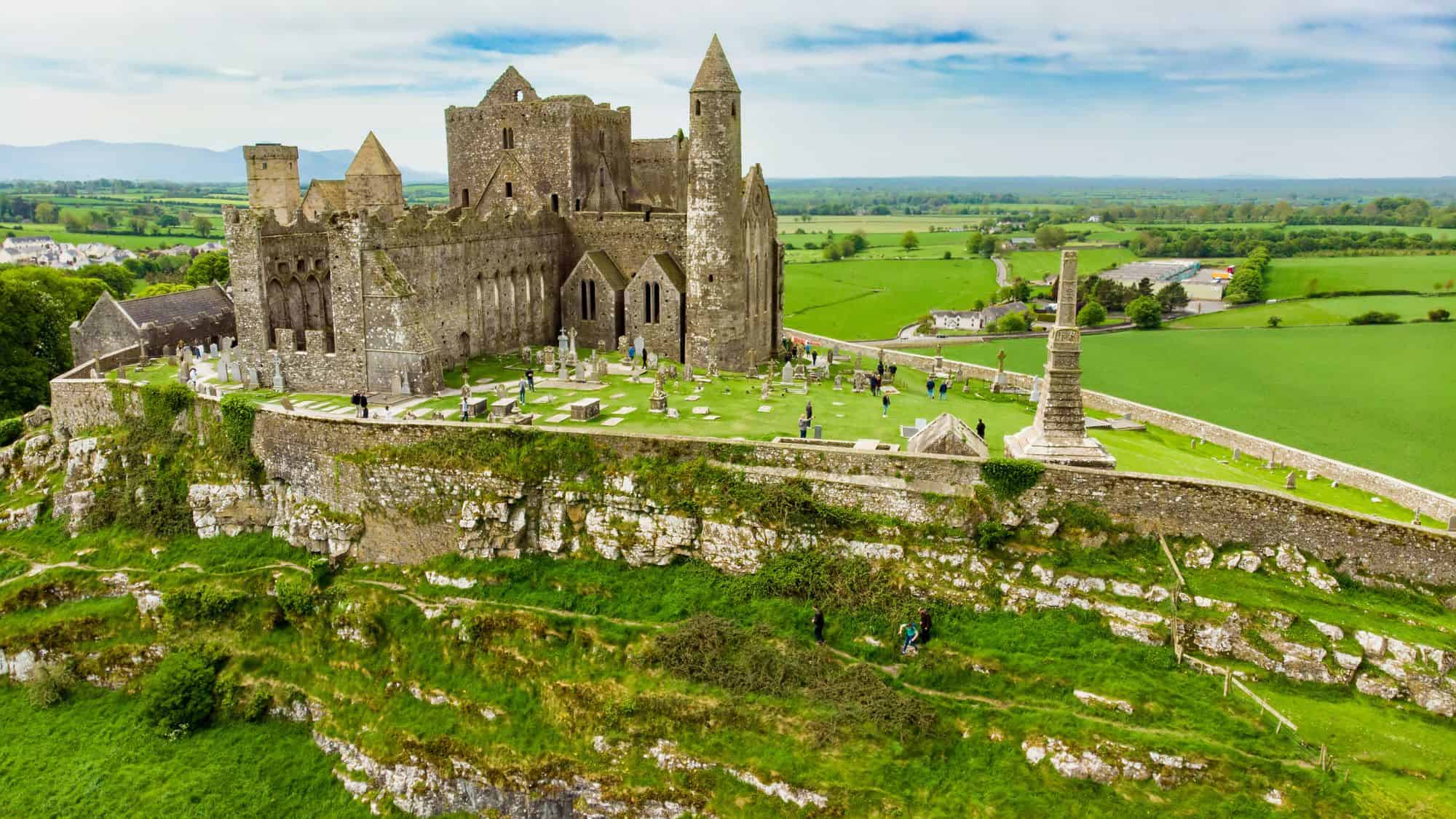 An aerial view of the Rock of Cashel in County Tipperary, Ireland, showing the medieval castle complex perched on a limestone hill. The surrounding vibrant green fields contrast with the gray stone of the historic site, with visitors exploring the grounds.
