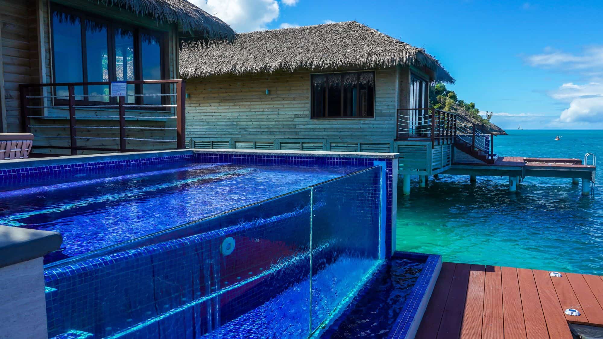 A luxurious overwater villa with a private infinity pool overlooking the turquoise sea. The villa has a modern design with a thatched roof and large glass windows for uninterrupted ocean views.