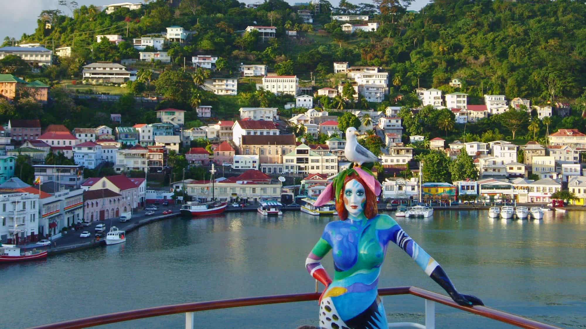A colorful painted statue of a woman with a seagull perched on her head stands on a balcony overlooking Saint George's harbor, with vibrant hillside houses and lush greenery in the background.
