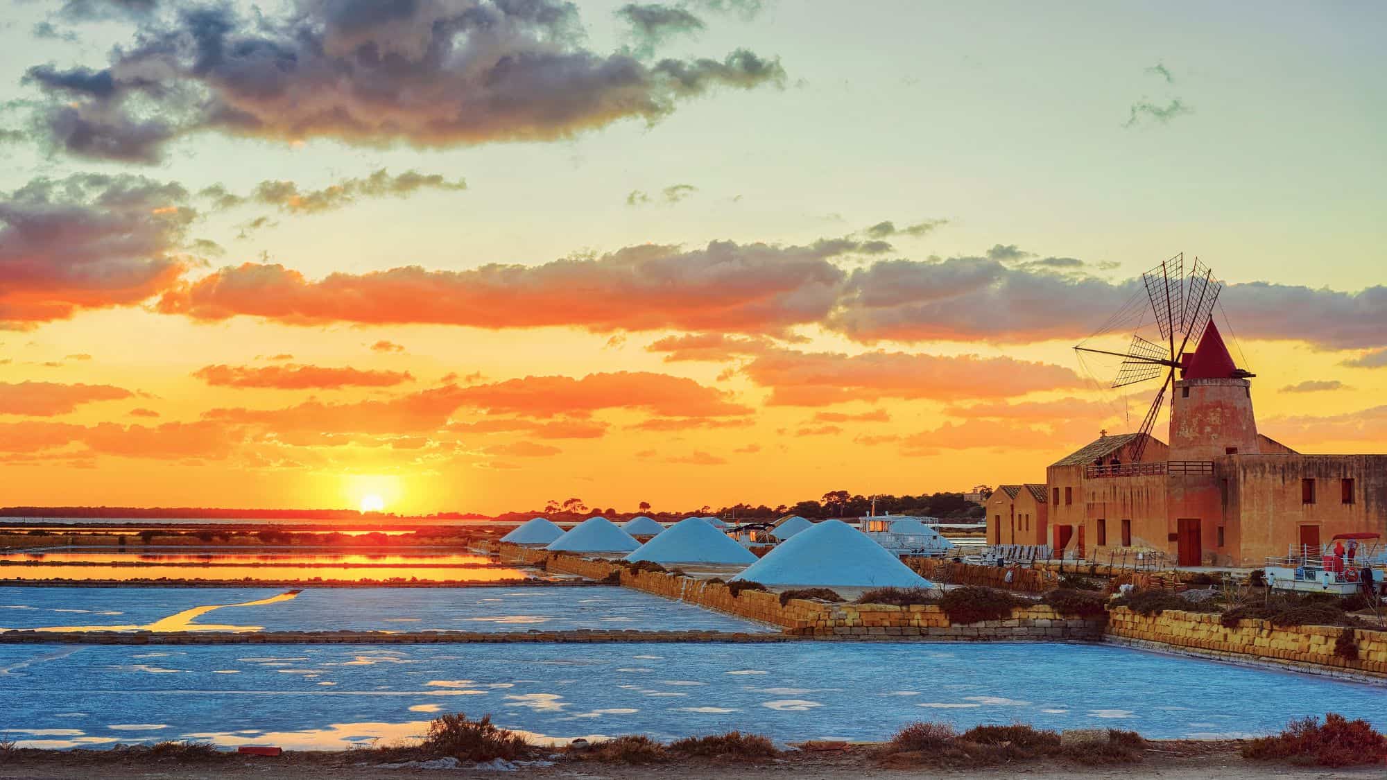 A vibrant sunset over the salt pans of Trapani, Sicily, with white salt mounds lined along reflective water channels. A traditional windmill stands as a historical reminder of the region's salt production heritage.