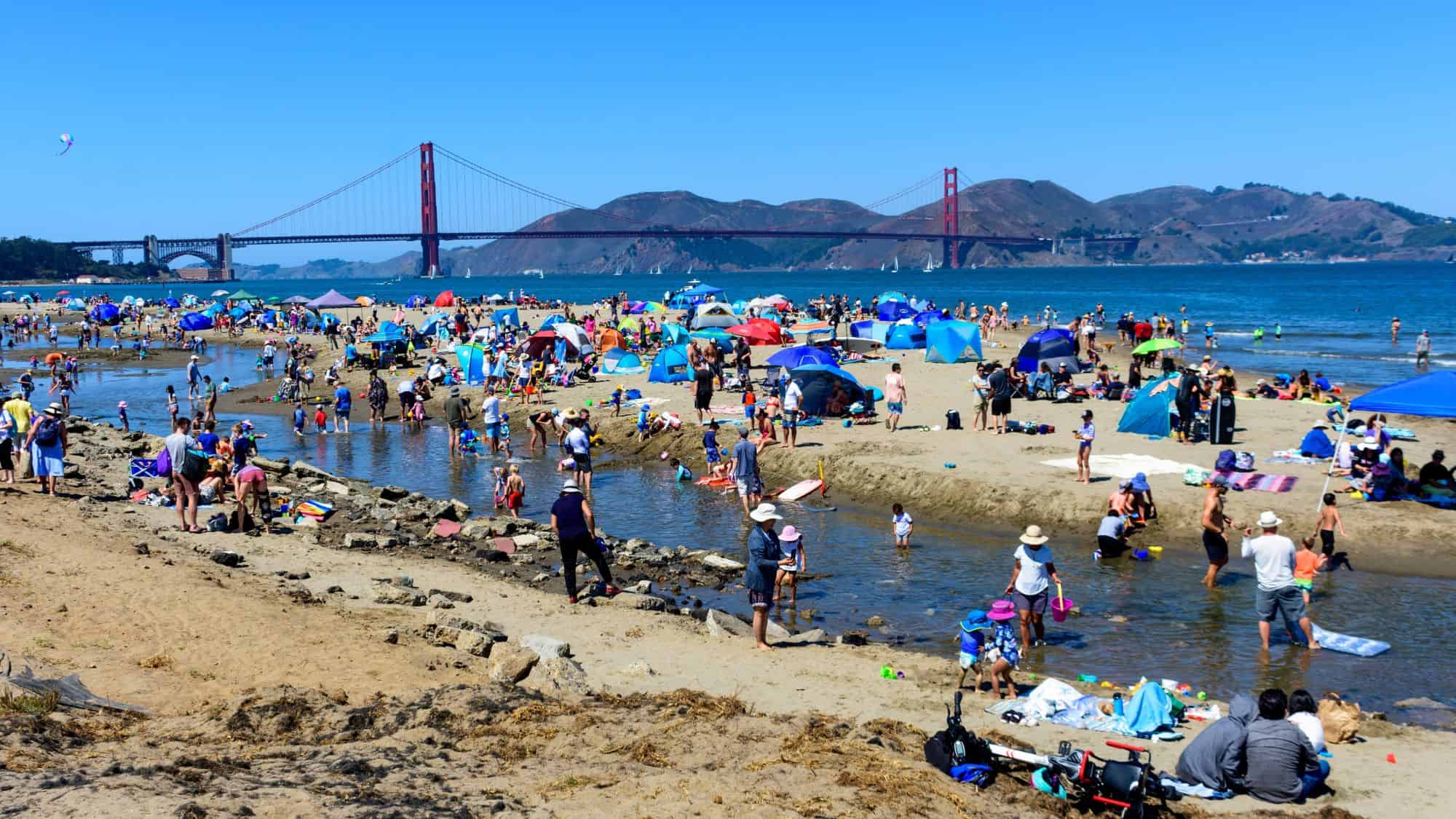 A lively beach scene in San Francisco, California, with people sunbathing, playing, and relaxing under colorful tents. The Golden Gate Bridge stands in the background, contrasting against the bright blue sky and rolling hills.