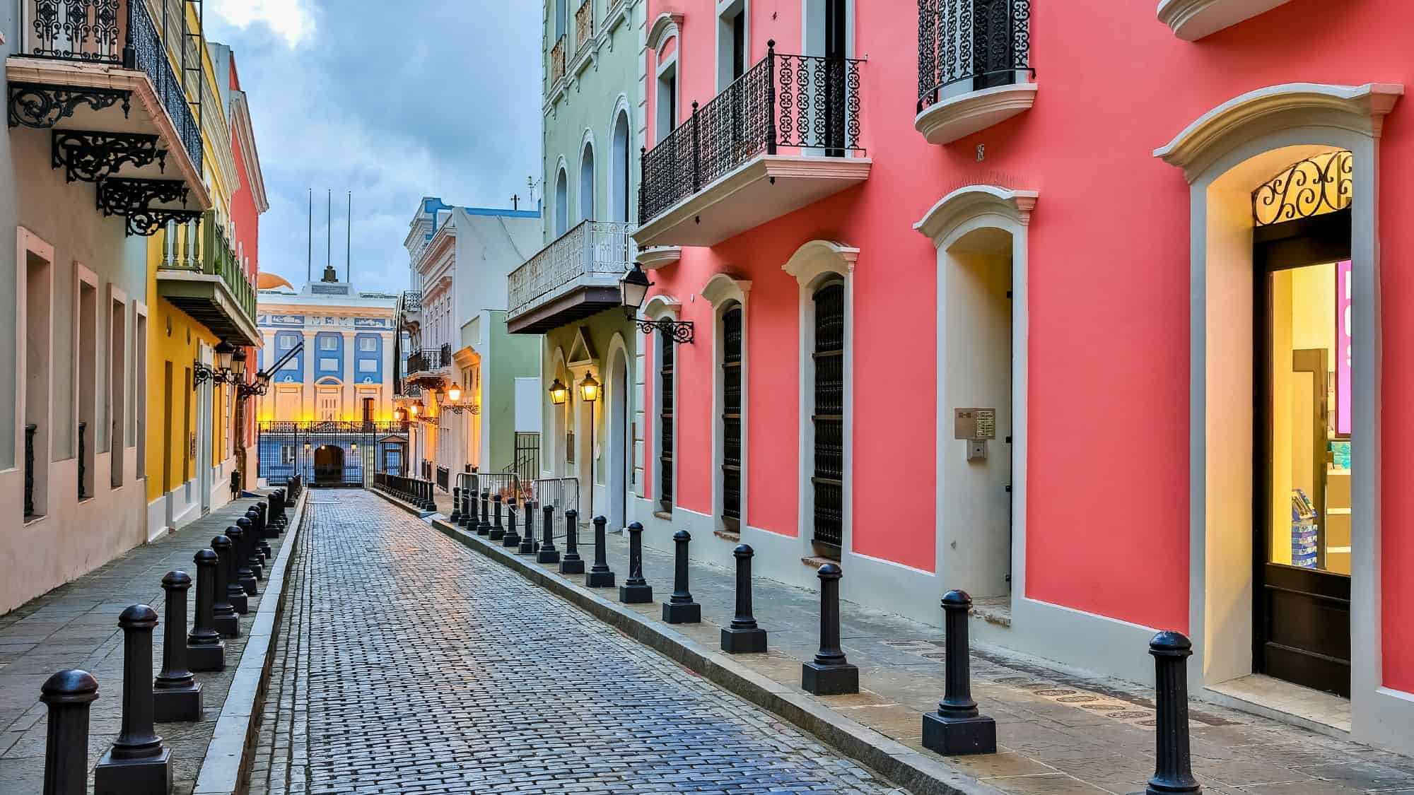 A charming cobblestone street in Old San Juan, Puerto Rico, is lined with colorful colonial buildings featuring wrought-iron balconies, leading toward a softly lit plaza at dusk.
