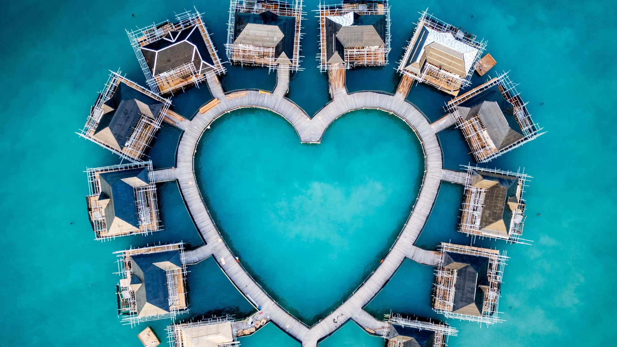 A heart-shaped arrangement of overwater bungalows connected by wooden walkways, floating on vibrant turquoise water. The aerial view captures the unique, romantic design of the resort.