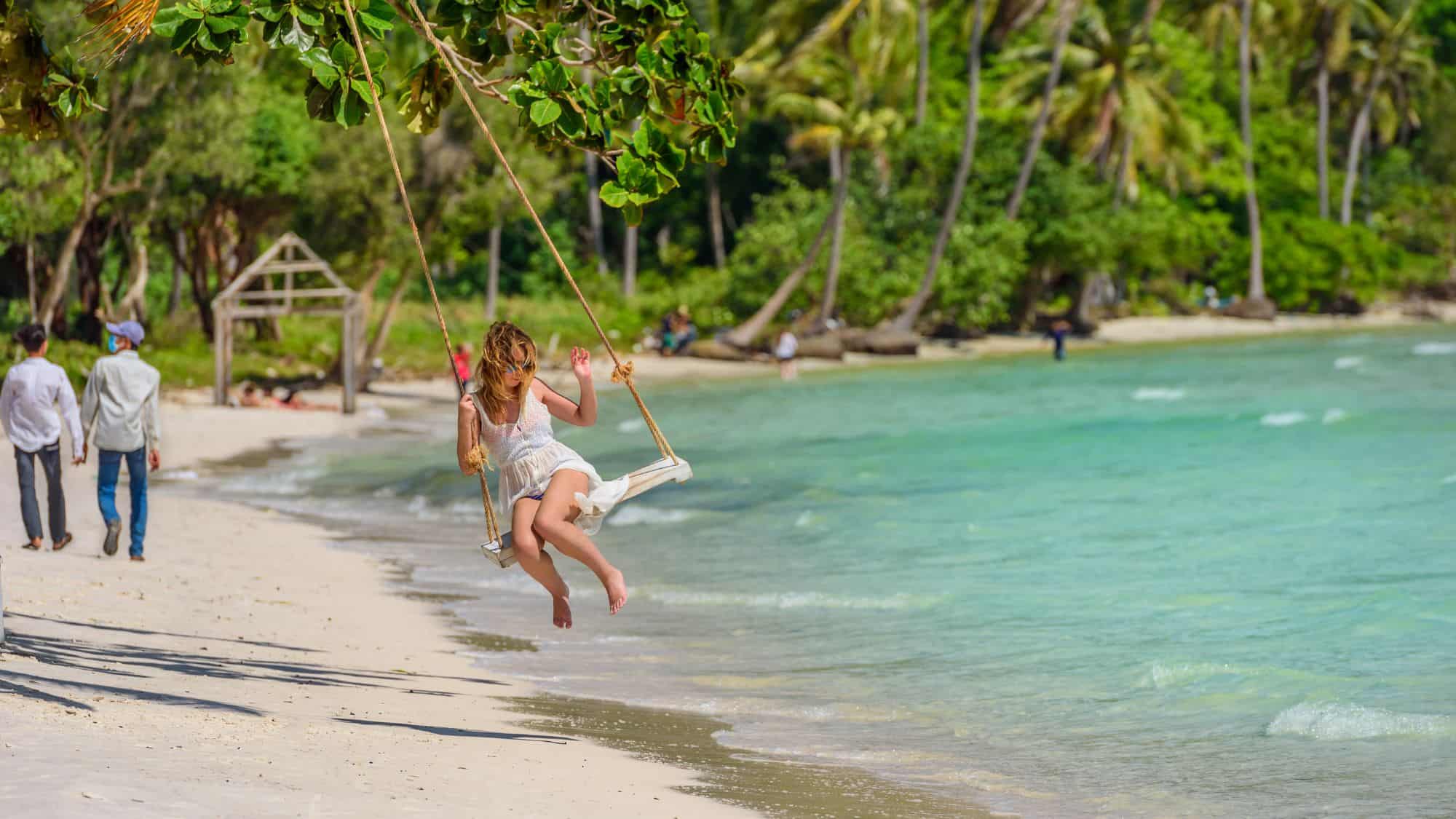 A dreamy tropical beach where a young woman in a white dress swings from a tree over the soft white sand, with turquoise waves gently lapping the shore.