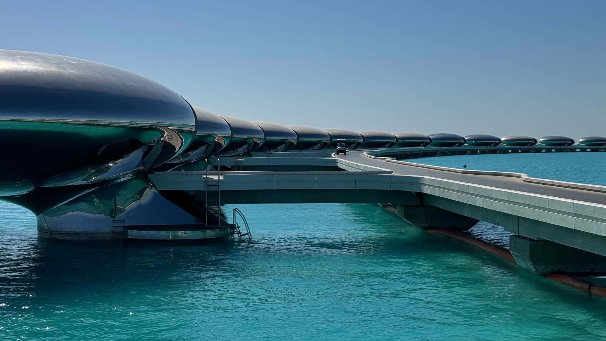 A futuristic, sleek resort with black, pod-like overwater villas connected by a smooth, curving walkway. The contrast between the modern architecture and the vivid turquoise water is striking.