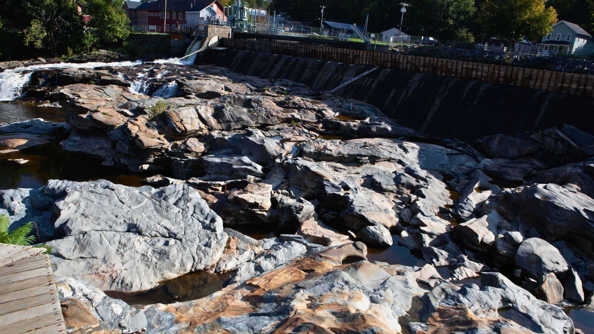Rock formations and potholes shaped by natural erosion are visible at Shelburne Falls, with water trickling through the crevices. A small dam and buildings from the nearby town can be seen in the background.