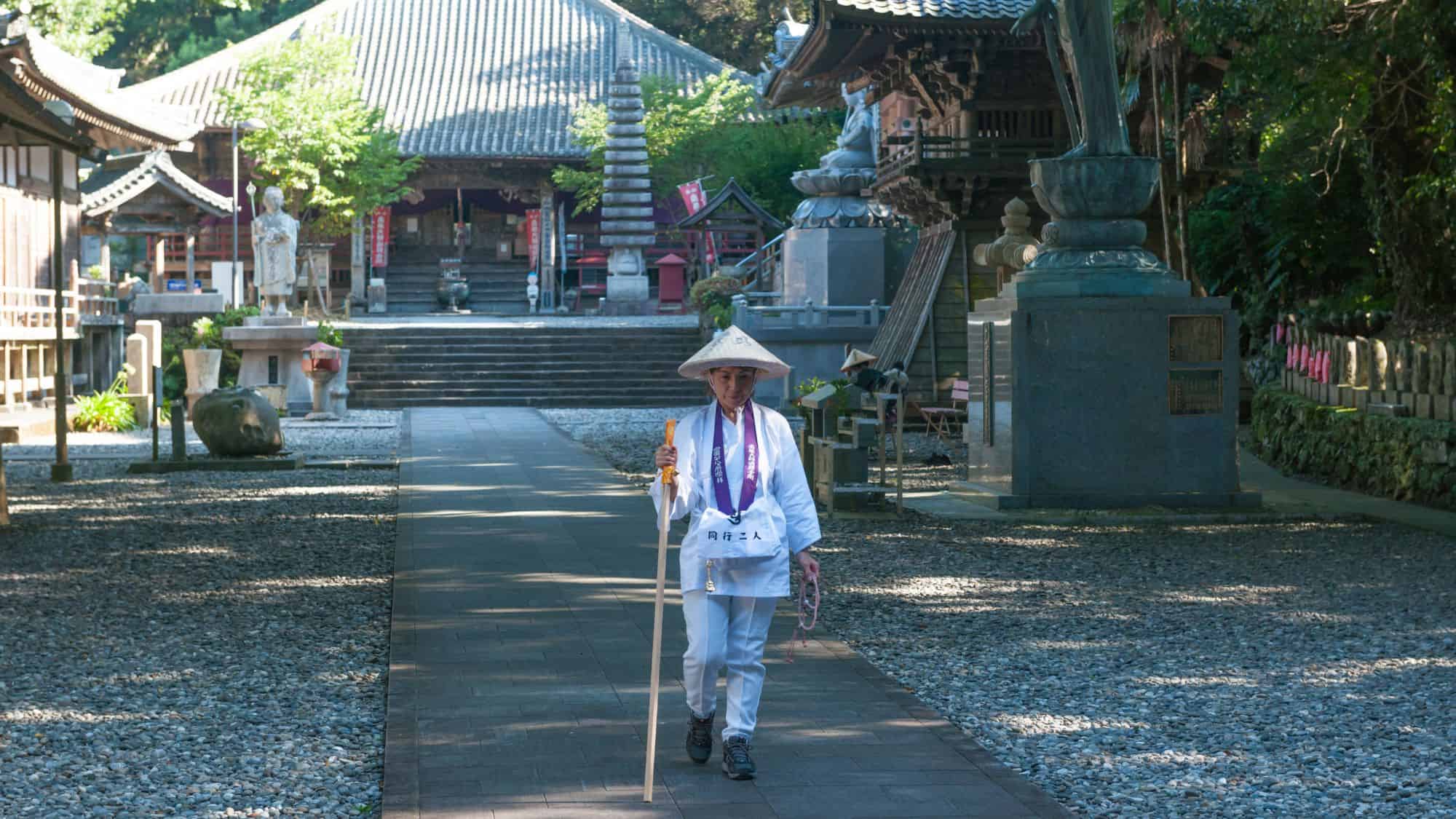 An elderly pilgrim in traditional white attire walks along a paved path leading to a Japanese temple. The temple grounds include stone lanterns, a statue, and lush greenery surrounding the historical structures.