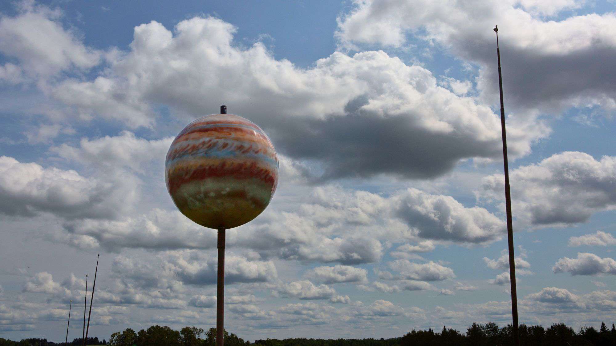 A large, painted model of Jupiter mounted on a pole stands under a partly cloudy sky. The surrounding landscape features open fields and trees, adding a quirky roadside attraction vibe to the scene.