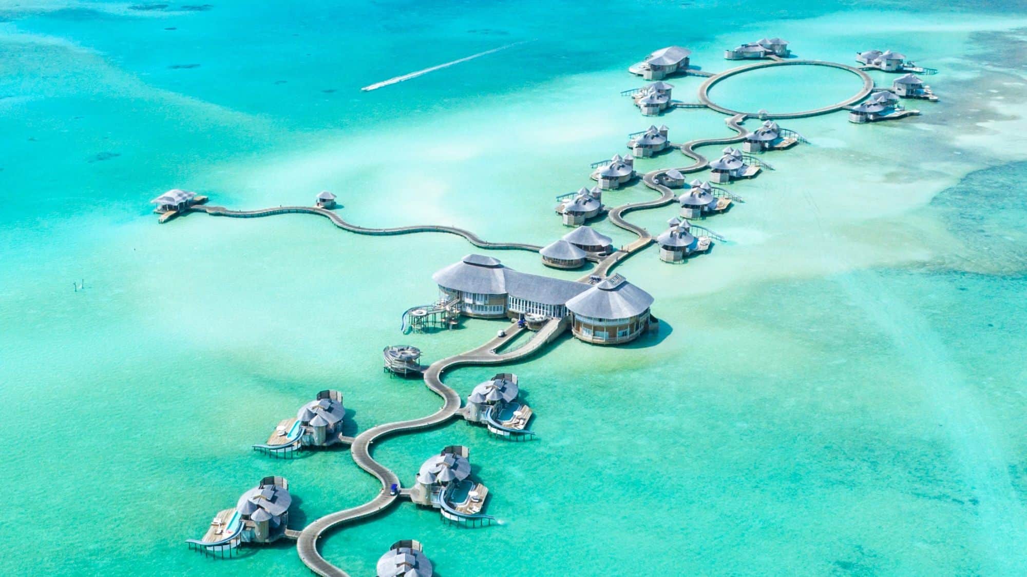 Aerial view of luxurious overwater villas with private pools, connected by winding boardwalks, set against a shallow turquoise lagoon. The villas are spaced out for privacy, with some clustered in a circular formation.