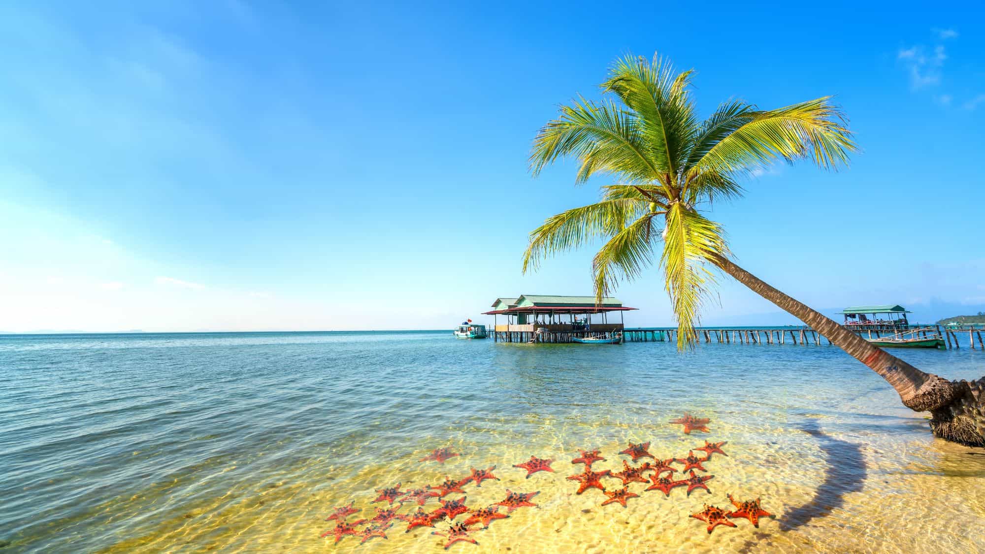 A serene beach scene with clear blue waters, rustic fishing boats, and weathered wooden structures along the shore, with distant islands visible on the horizon.