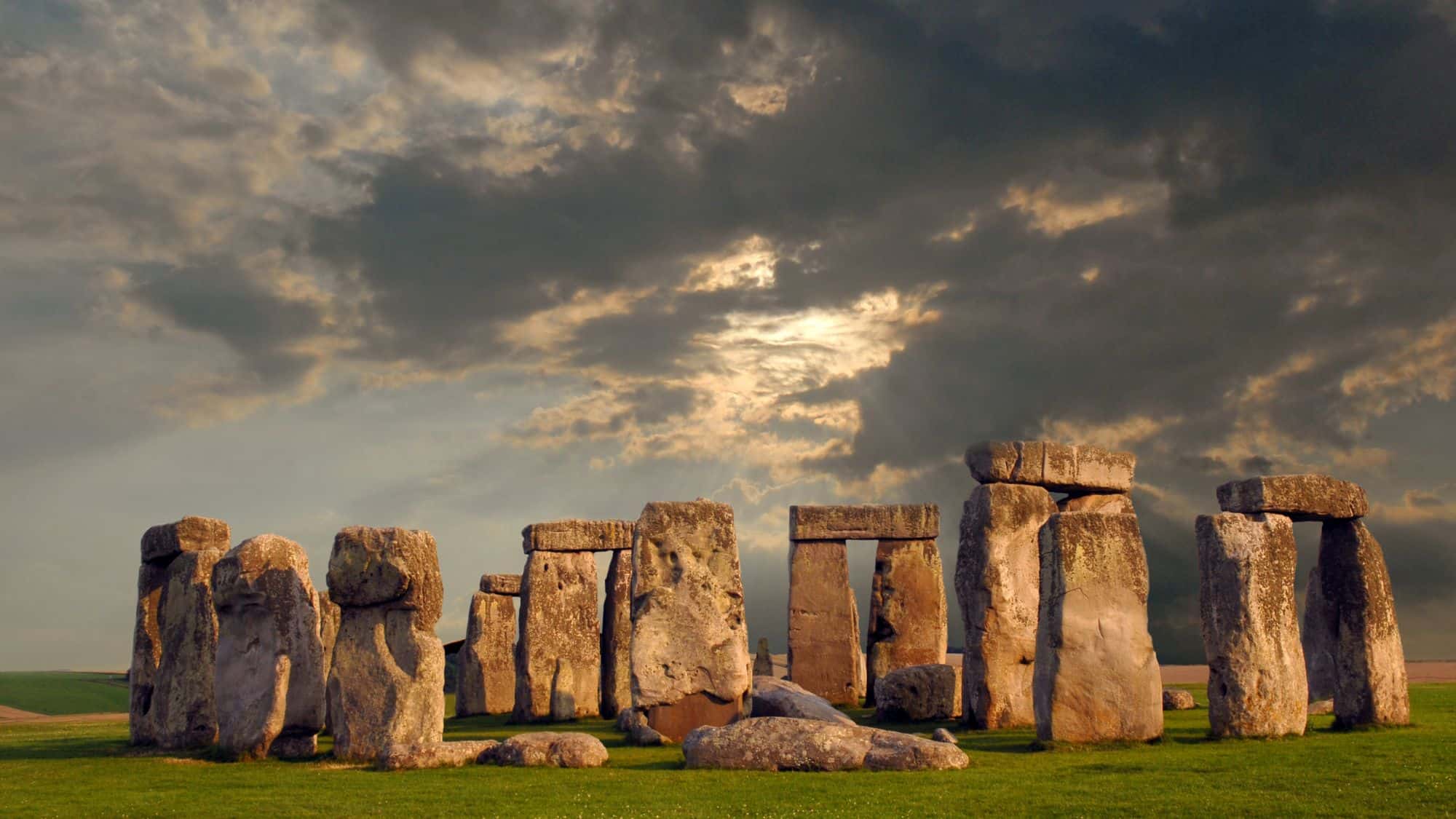 A dramatic view of Stonehenge, the ancient prehistoric stone circle in Wiltshire, England, set against a moody sky with dark clouds and a break of sunlight. The massive, weathered stones stand tall on a lush green plain.