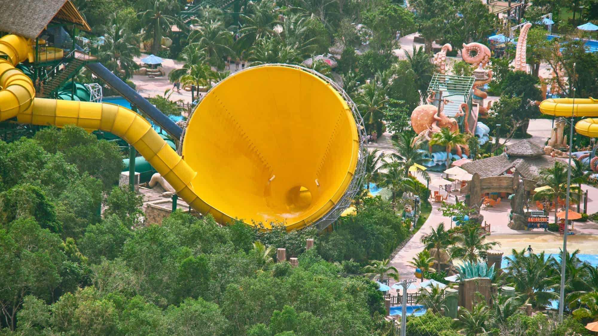 A thrilling aerial view of a tropical water park featuring a massive yellow waterslide shaped like a funnel, surrounded by lush greenery, palm trees, and various themed attractions.