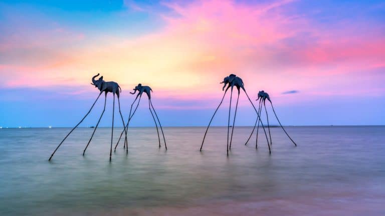A surreal sunset over the ocean featuring artistic long-legged elephant sculptures standing in the water, silhouetted against the pastel hues of the sky.