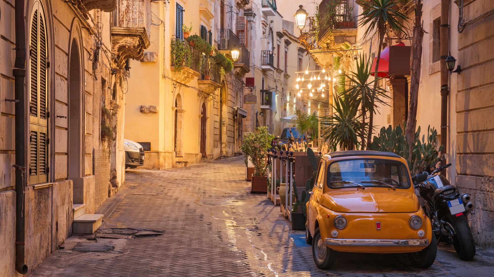 A charming street in Syracuse, Italy, with warm-toned historic buildings, potted plants, and string lights. A vintage yellow Fiat parked alongside a motorbike enhances the nostalgic, cozy atmosphere.