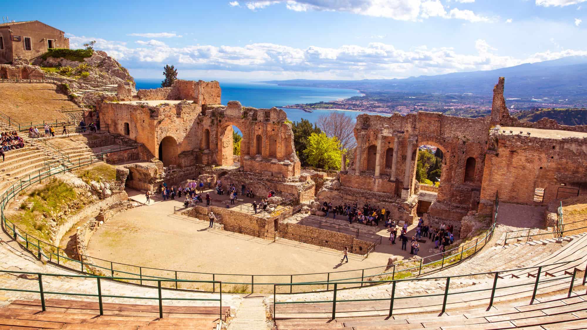 The ancient Greek-Roman theater in Taormina, Sicily, with its stone arches and seating area overlooking the sea and Mount Etna. Tourists explore the historic site under a sunny sky.