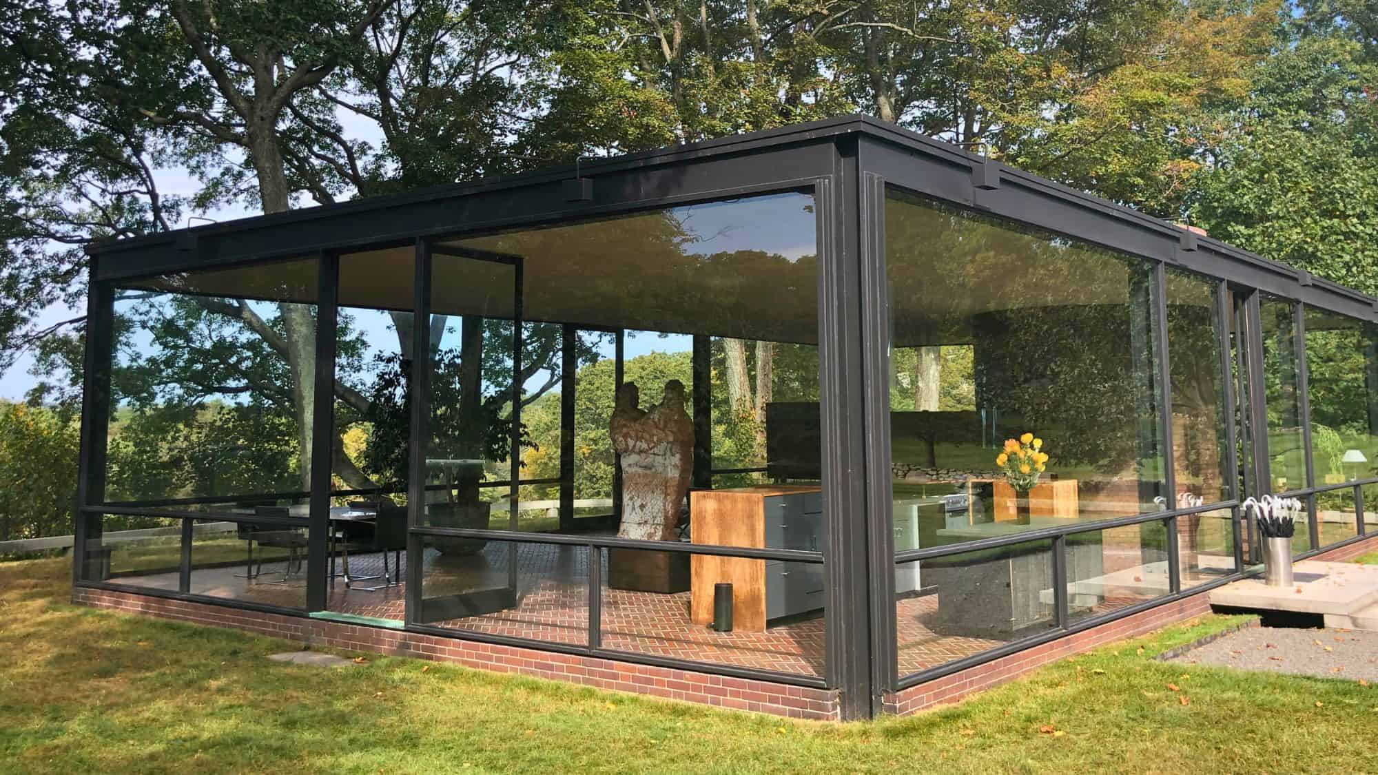A modern, minimalist glass house with floor-to-ceiling windows sits amidst a wooded landscape. The transparent walls reveal sleek interior furnishings, blending architecture with nature seamlessly.