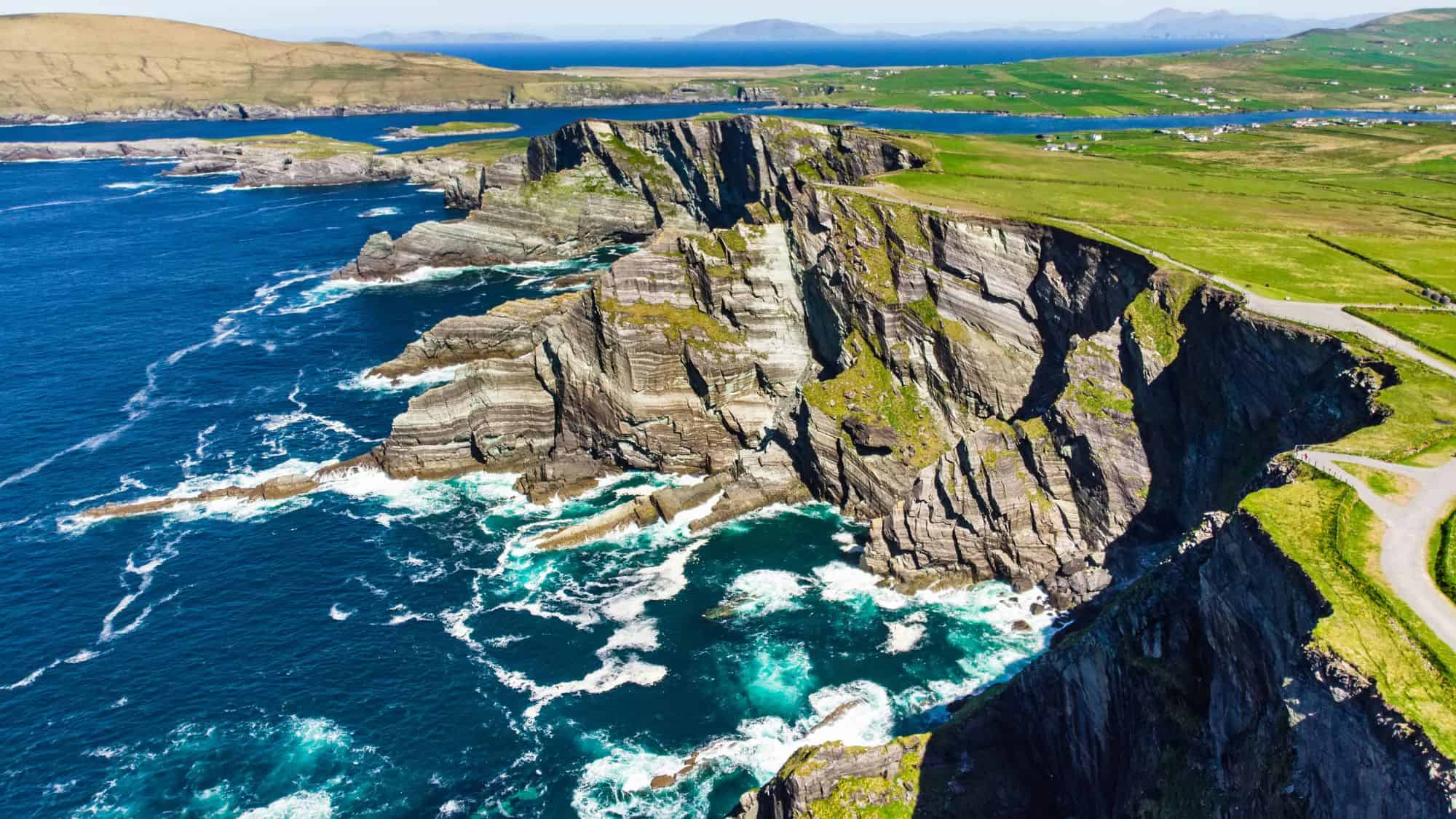 A stunning aerial view of the Kerry Cliffs along Ireland's Wild Atlantic Way. Jagged cliffs rise from the crashing turquoise waves, with rolling green hills and winding coastal roads in the background.
