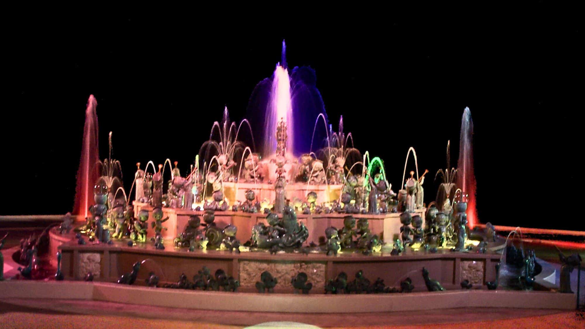 A colorful and intricately designed water fountain featuring multiple small statues, illuminated with vibrant lighting at night. The display showcases a whimsical and artistic theme with water jets shooting in various directions.