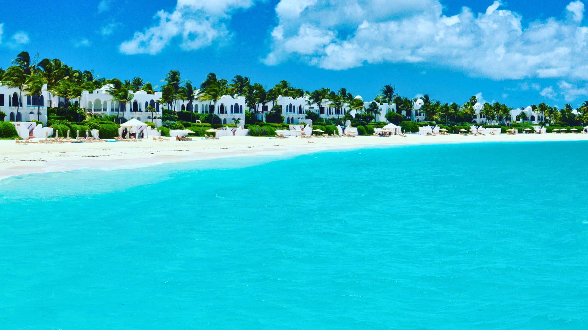 A luxurious stretch of white sand beach with clear, bright blue waters. Elegant white beachfront villas with arched windows are surrounded by lush greenery and palm trees.