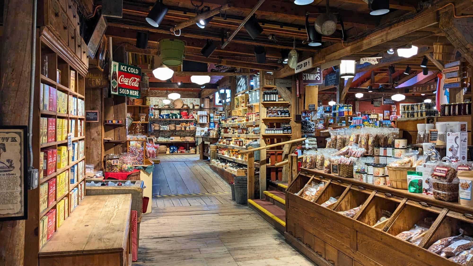 A cozy, rustic interior of The Vermont Country Store features wooden shelves lined with colorful packaged goods, baskets of snacks, and vintage signs, including one for Coca-Cola. The wooden beams and soft lighting add to its nostalgic charm.
