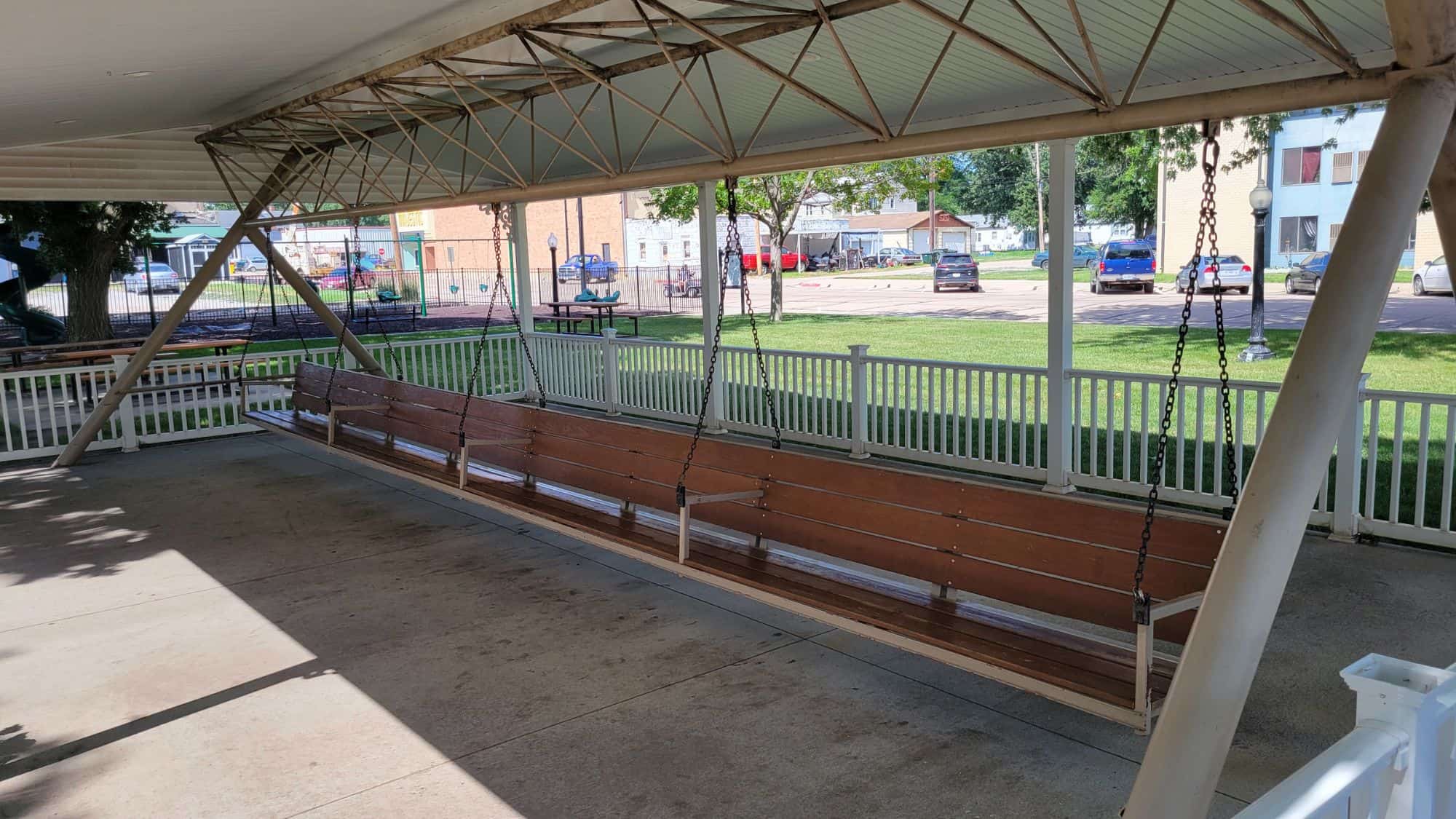 An enormous wooden porch swing, suspended from a covered pavilion, stretches across the seating area. The simple wooden swing is part of a community gathering spot, surrounded by a white fence with a small-town setting in the background.