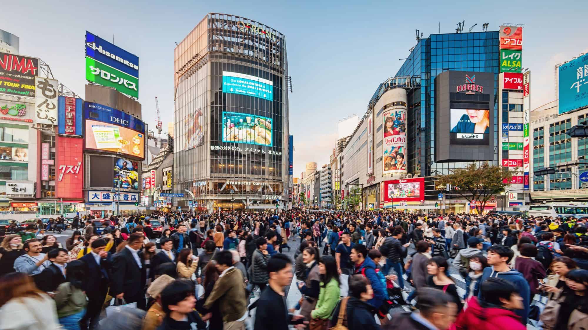 A bustling view of Shibuya Crossing in Tokyo, Japan, filled with pedestrians crossing in all directions amid towering buildings covered in bright advertisements and billboards. The famous urban intersection is surrounded by a mix of modern and traditional architecture.