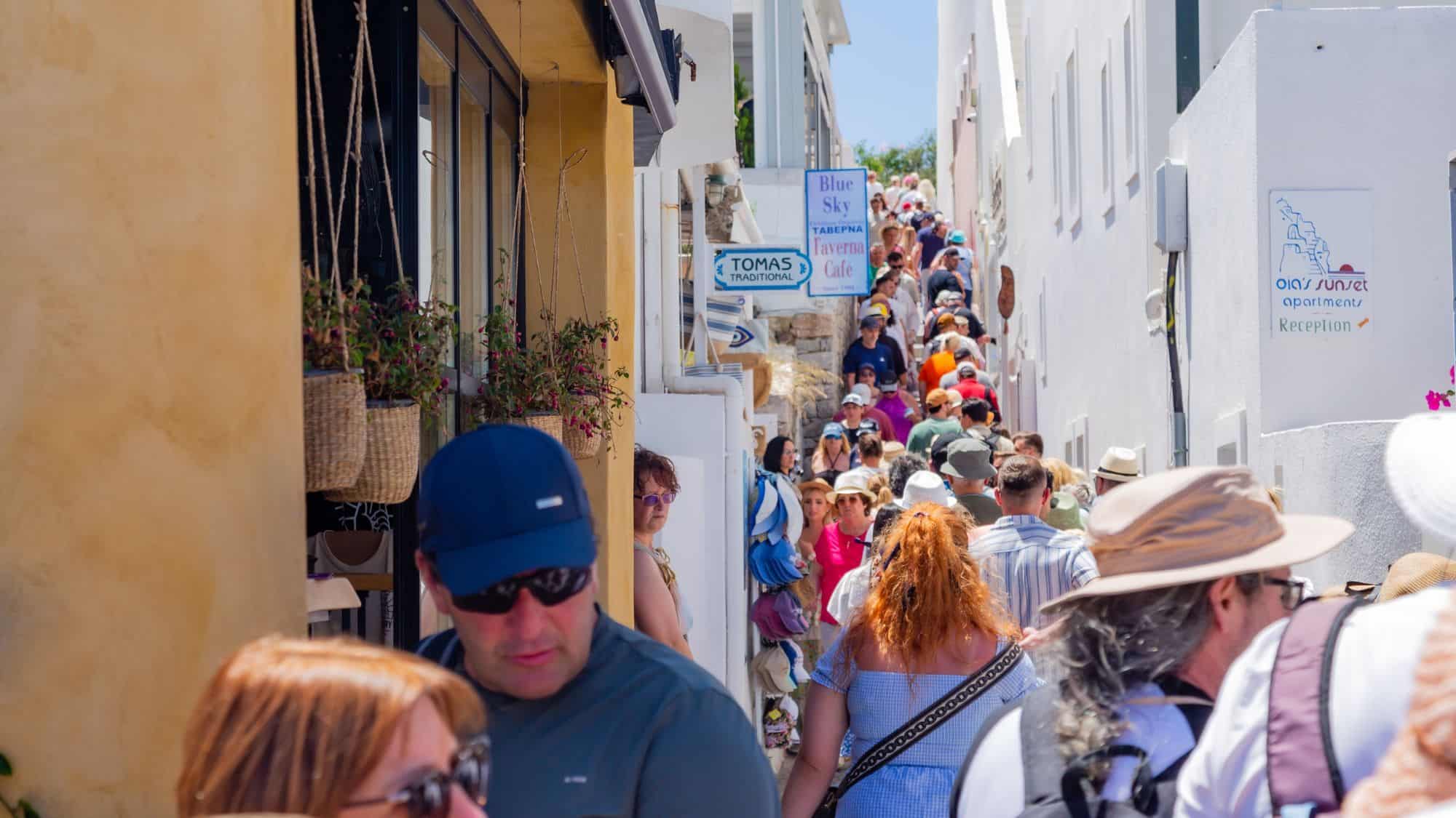 A steep, narrow alley in Santorini, Greece, crowded with tourists in sun hats and backpacks navigating through the white-washed buildings. The signs for local shops and taverns add to the charming Mediterranean atmosphere.