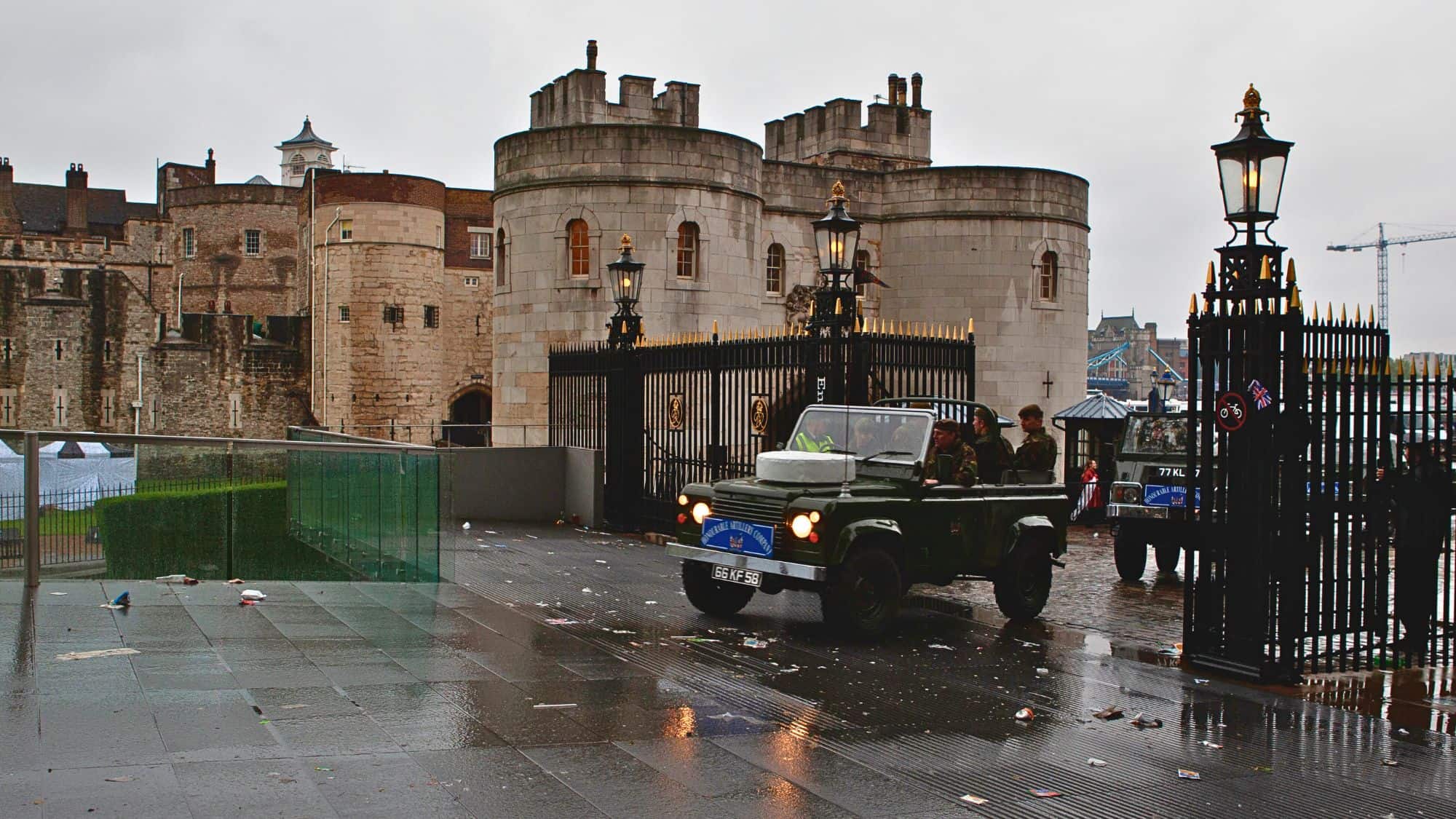 The historic Tower of London, with its stone walls and turrets, stands behind black iron gates. Military vehicles are parked at the entrance, and the wet ground reflects the cloudy sky.
