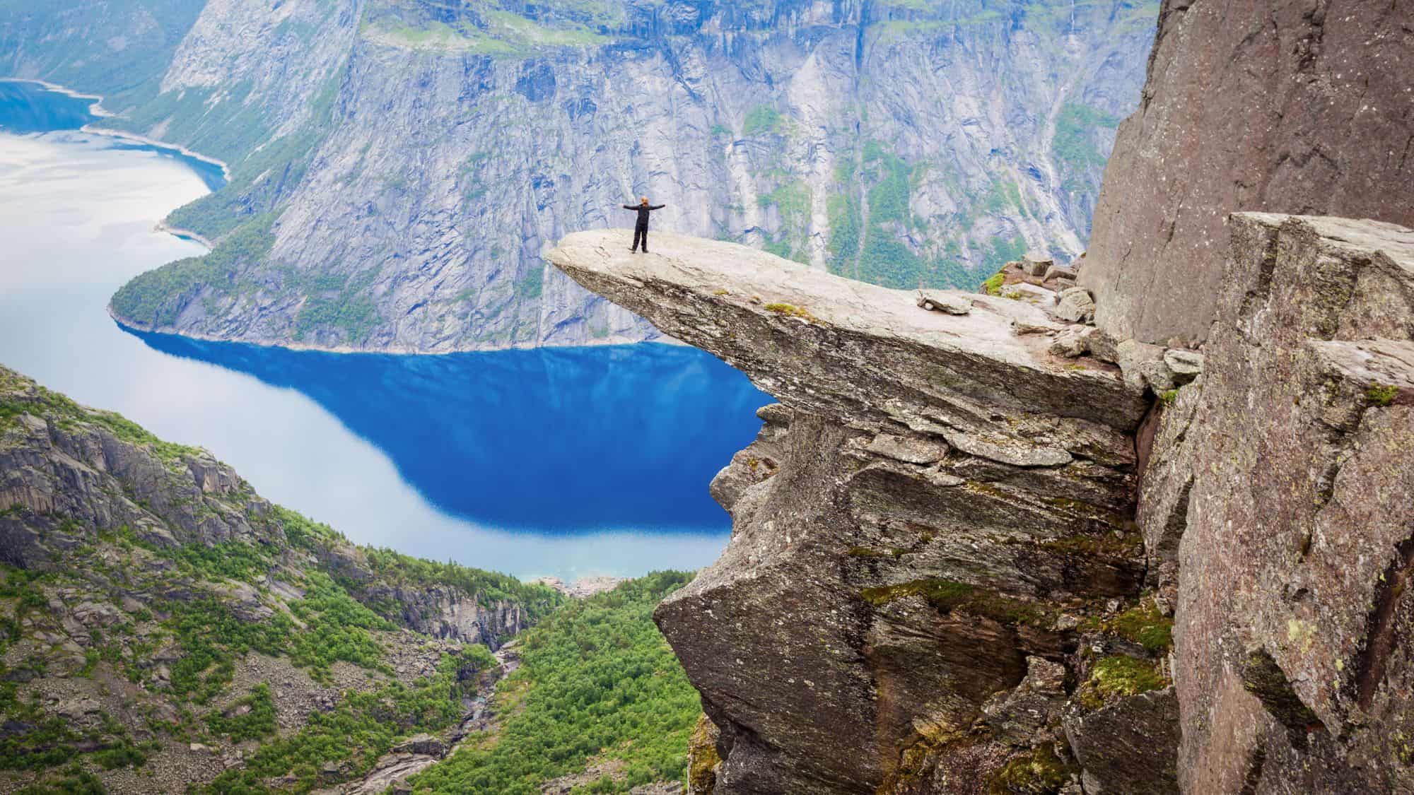 A breathtaking scene of the iconic Trolltunga rock formation jutting out over a deep blue fjord, with a hiker standing triumphantly at the edge, surrounded by towering cliffs and lush greenery.
