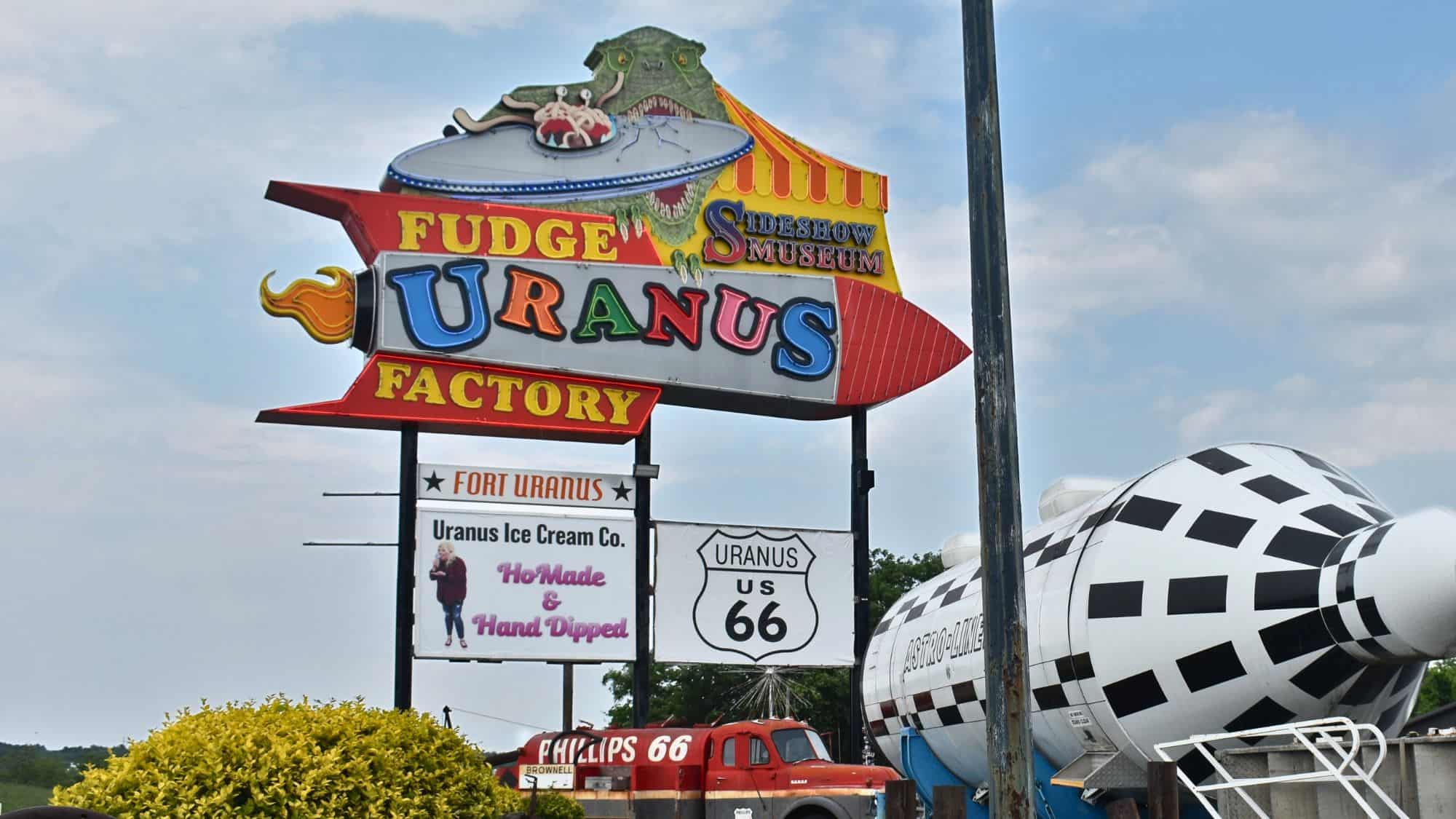 A bold and playful sign for “Uranus Fudge Factory,” featuring bright neon colors, a rocket ship design, and additional signage for the sideshow museum. The location is a humorous and well-known stop along Route 66.