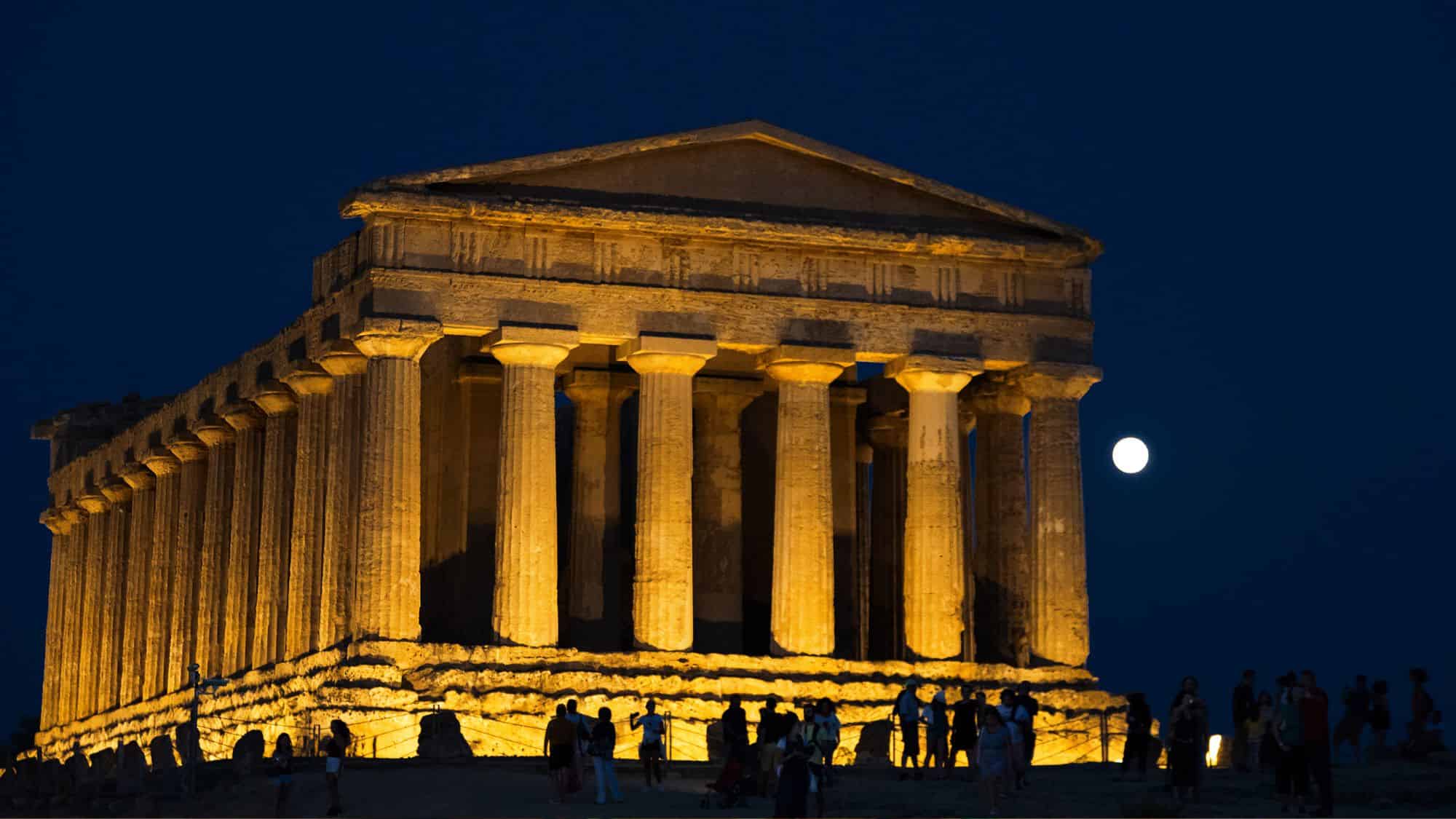 The Temple of Concordia in the Valley of the Temples, Agrigento, illuminated against the dark night sky. The ancient Greek structure glows with golden light, while visitors explore the site under the moonlight.