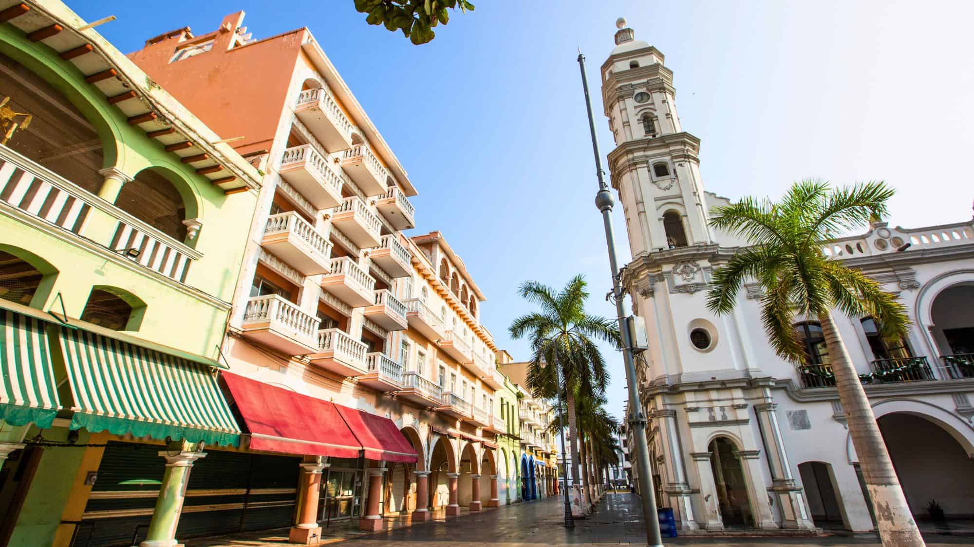 A picturesque street in Veracruz, Mexico, featuring colonial-style buildings with colorful facades, arched walkways, and a historic white church tower. Palm trees line the street, and the bright blue sky adds to the lively atmosphere.