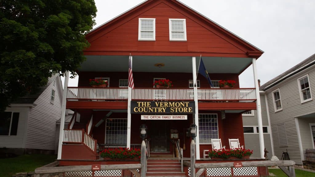 A charming red clapboard building houses The Vermont Country Store, with an inviting front porch decorated with rocking chairs and flower boxes. The sign proudly states "The Orton Family Business," and two flags, including the American flag, are displayed at the entrance.