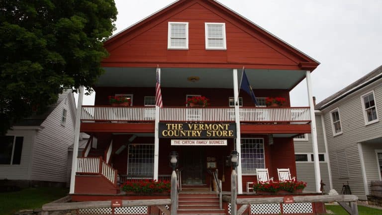 A charming red clapboard building houses The Vermont Country Store, with an inviting front porch decorated with rocking chairs and flower boxes. The sign proudly states "The Orton Family Business," and two flags, including the American flag, are displayed at the entrance.