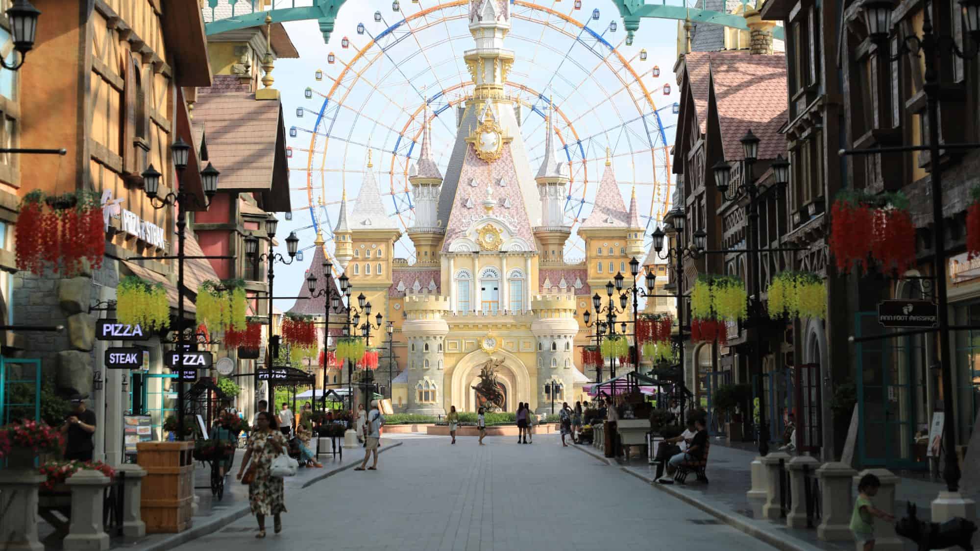 A fairy tale-like amusement park entrance with a grand castle, a giant Ferris wheel in the background, and a lively street lined with colorful hanging flowers, cafes, and shops.