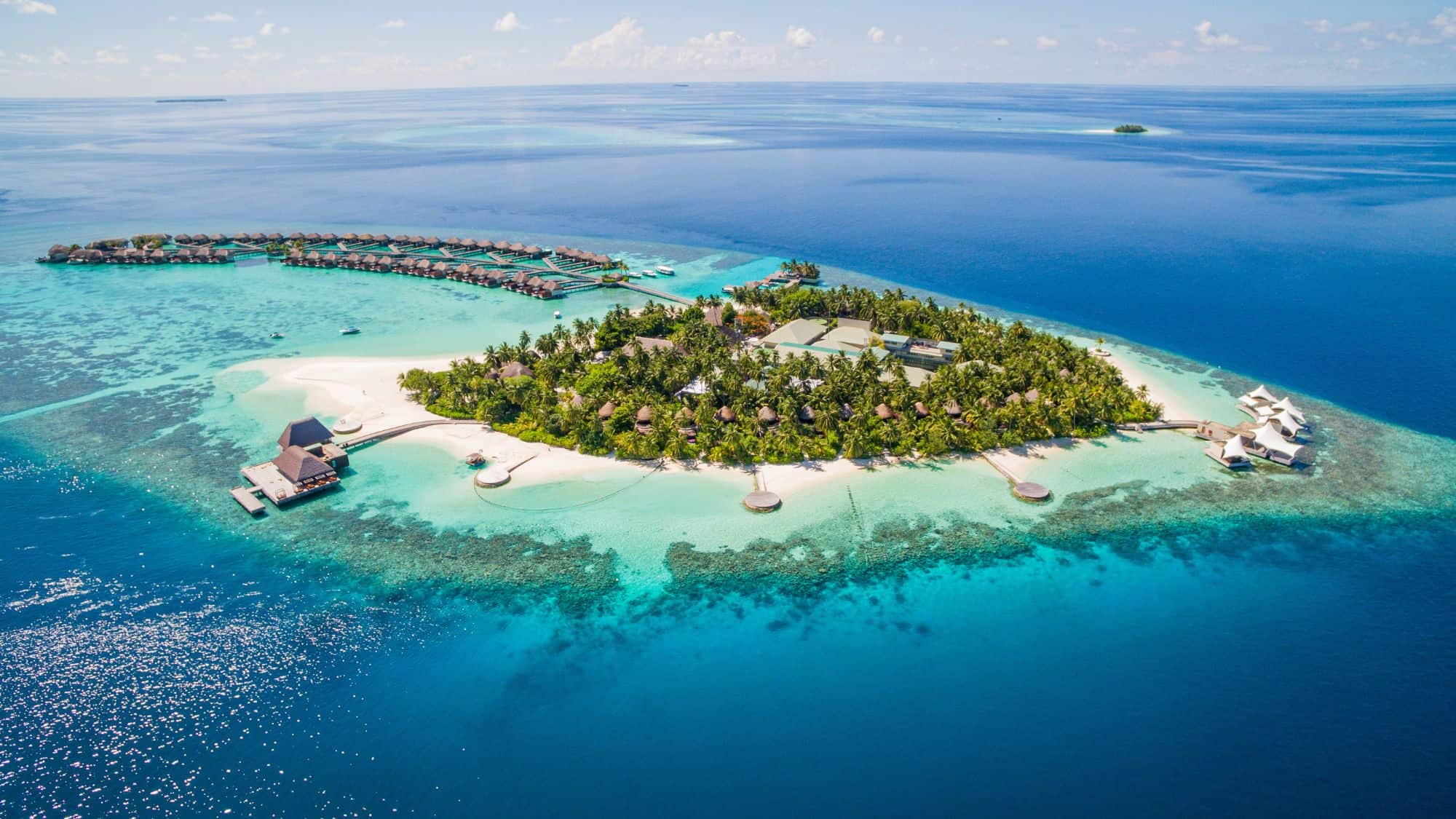 A stunning aerial view of a private island resort surrounded by turquoise waters, with overwater villas extending into the sea and lush greenery covering the island’s core.