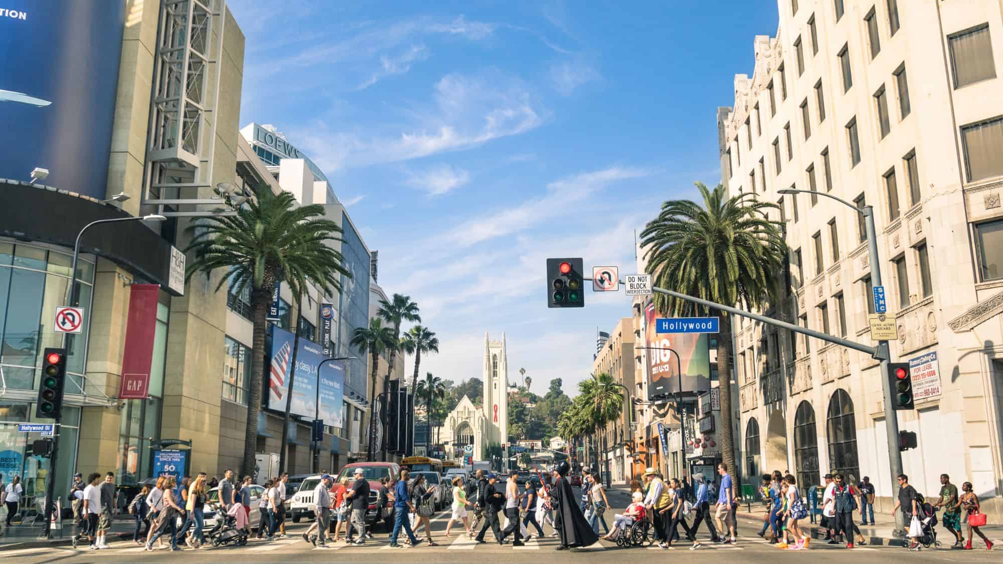 A busy street scene at the Hollywood Walk of Fame in Los Angeles, California, with tourists walking past palm trees, billboards, and famous landmarks. A costumed Darth Vader character stands among the crowd, adding to the entertainment-filled atmosphere.