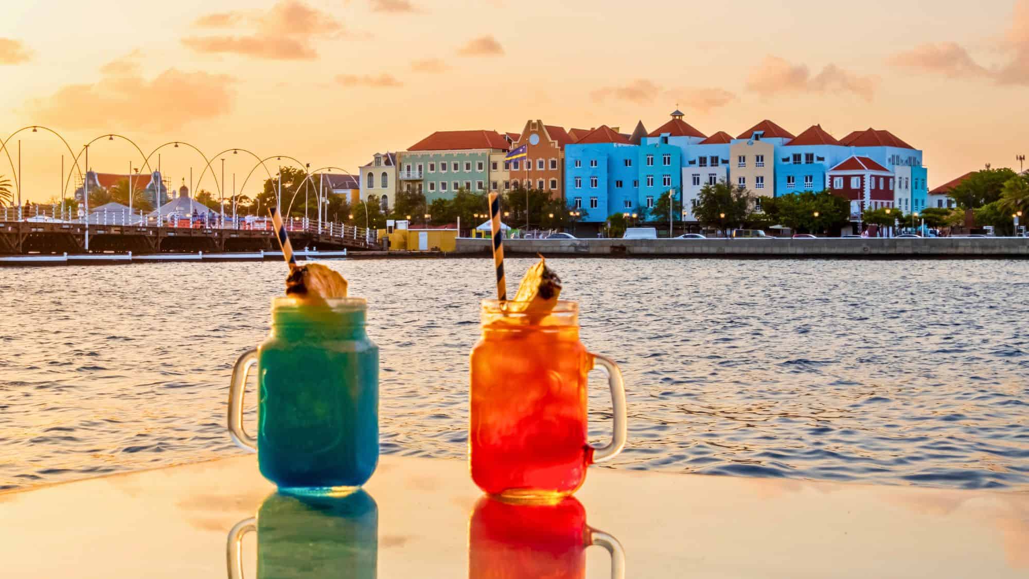 Two vibrant tropical drinks in mason jars sit on a table at sunset, with the iconic pastel-colored Dutch colonial buildings and the Queen Emma Bridge in the background.