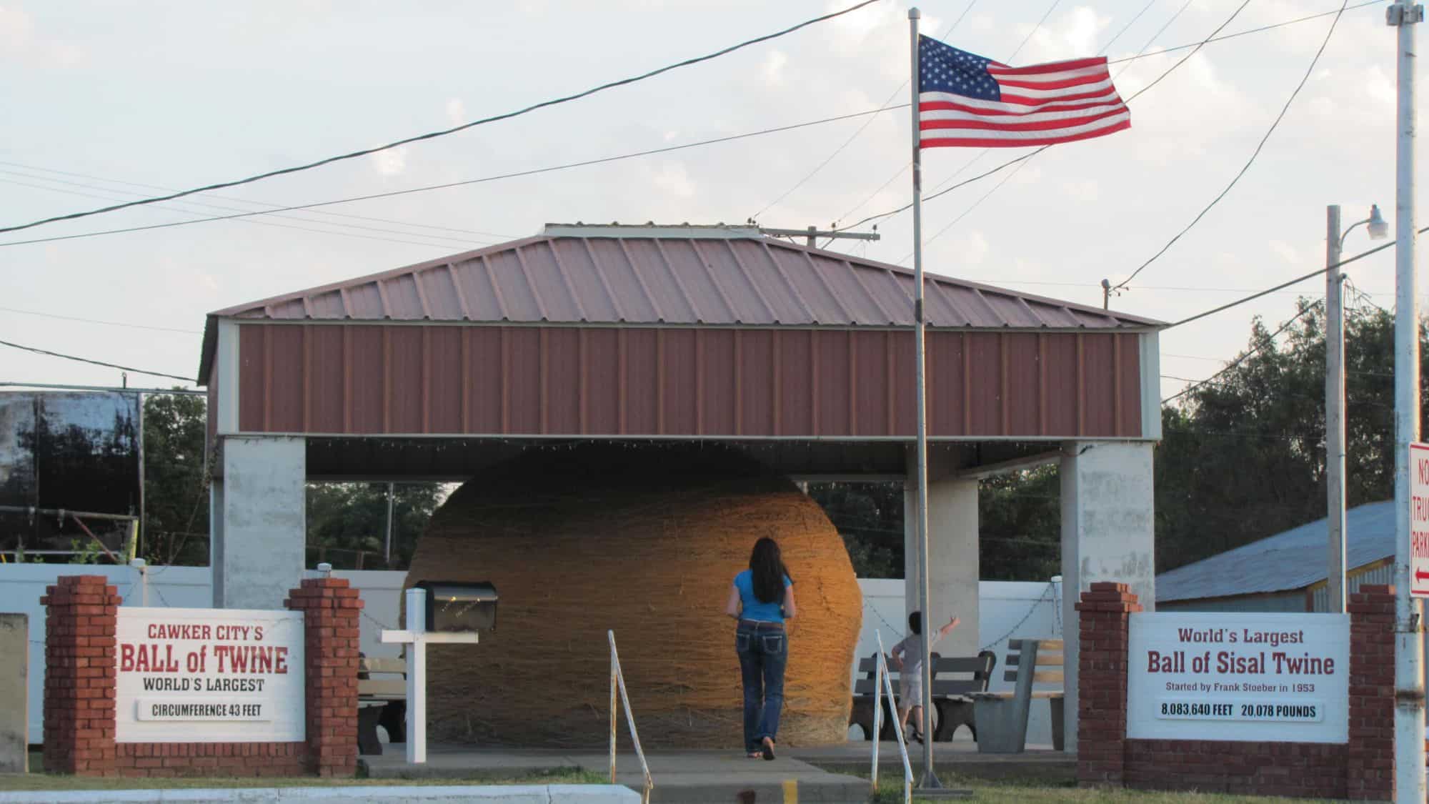A massive ball of sisal twine, housed under a metal pavilion, sits in Cawker City, Kansas. A sign reads "Cawker City's Ball of Twine - World's Largest," and an American flag flies above as a visitor approaches the enormous structure.